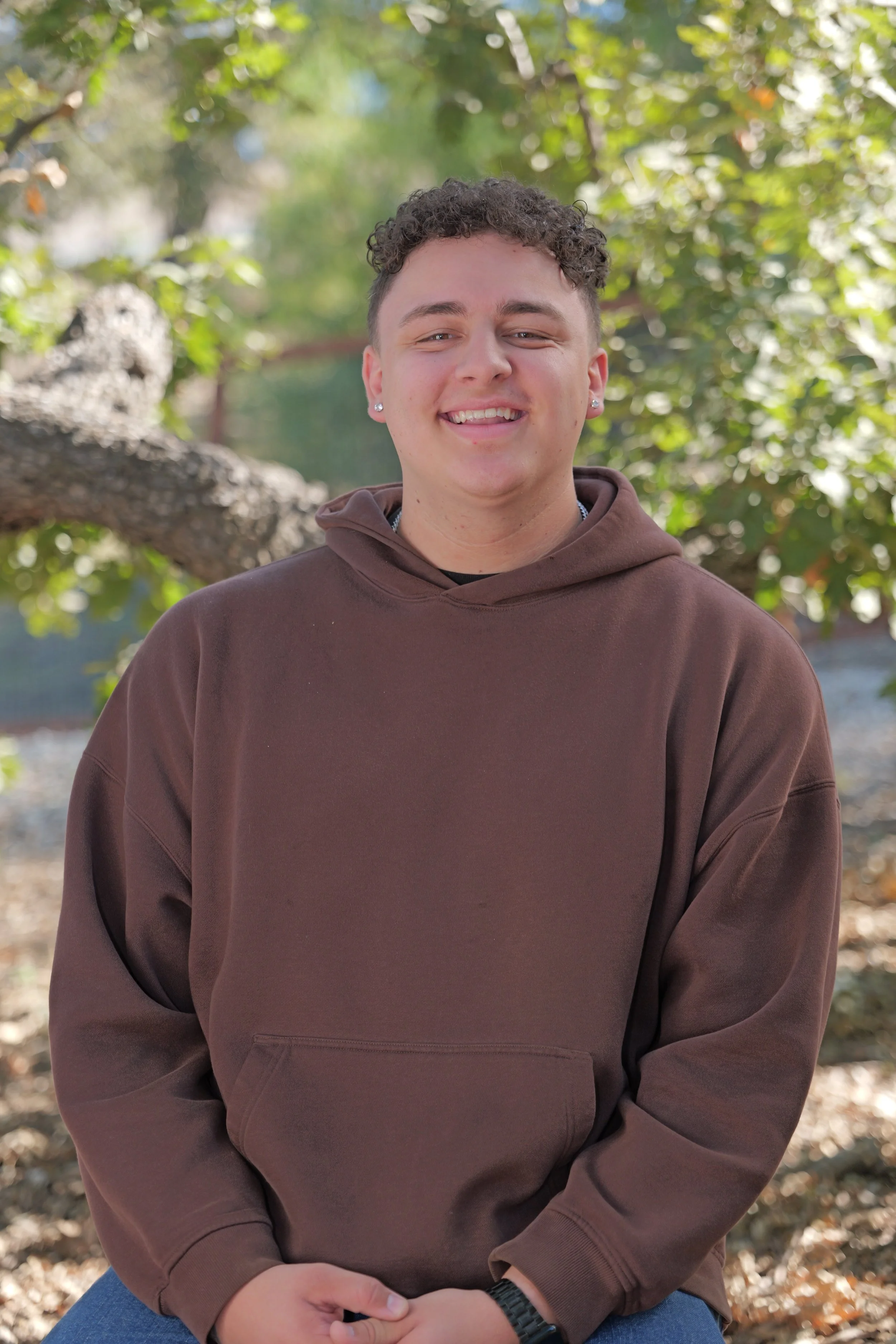 A young person smiling outdoors in a wooded area, wearing a black t-shirt, silver chain collar, earrings, and showing tattoos on their arms.