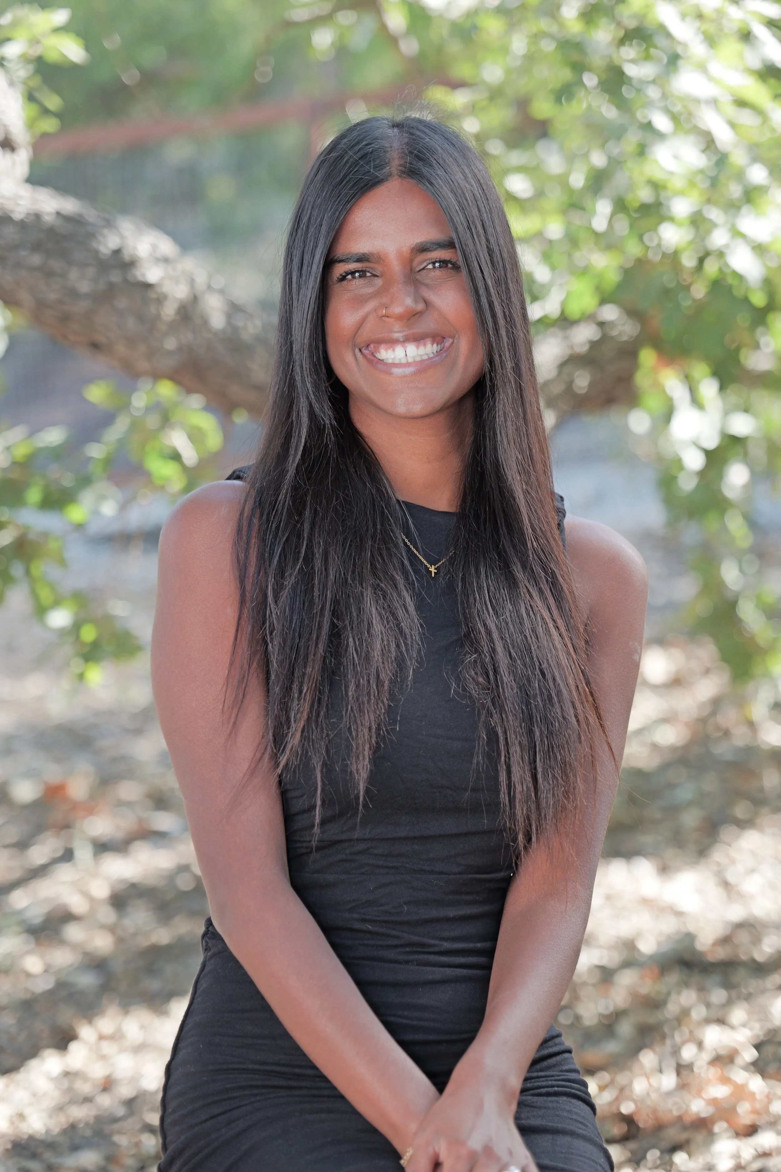 A woman with long dark hair and a nose piercing smiling outdoors in a natural setting.