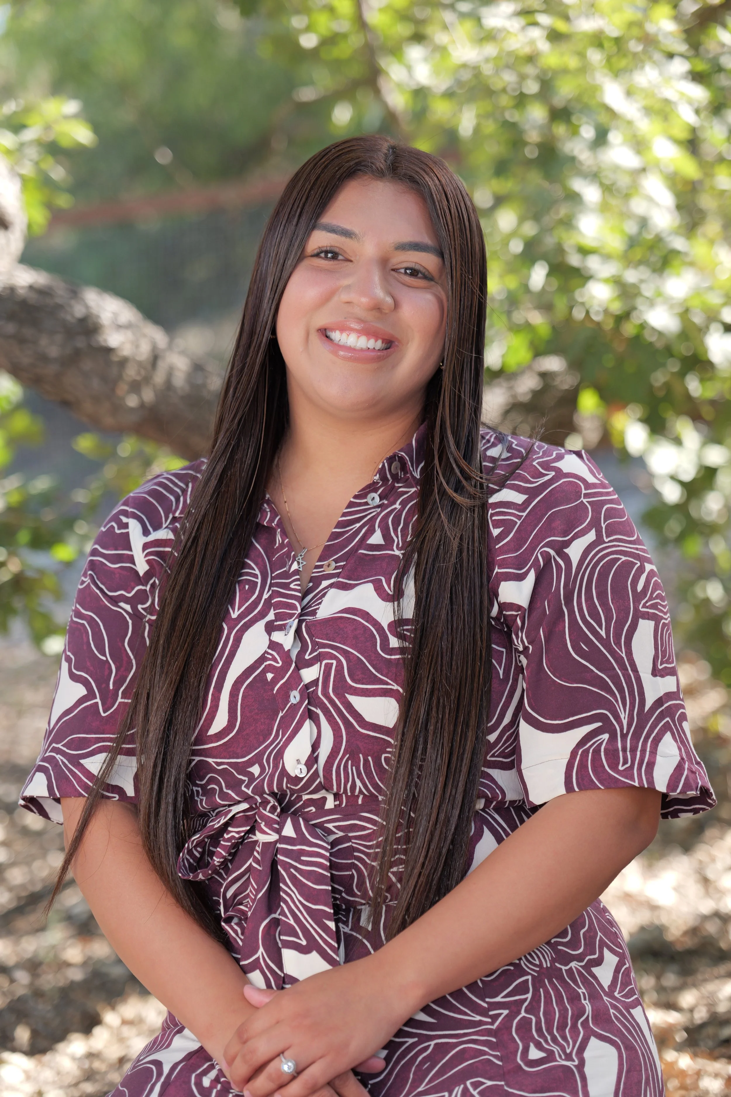 A woman with long brown hair, smiling, wearing a white blouse, outdoors with blurred trees and sunlight in the background.