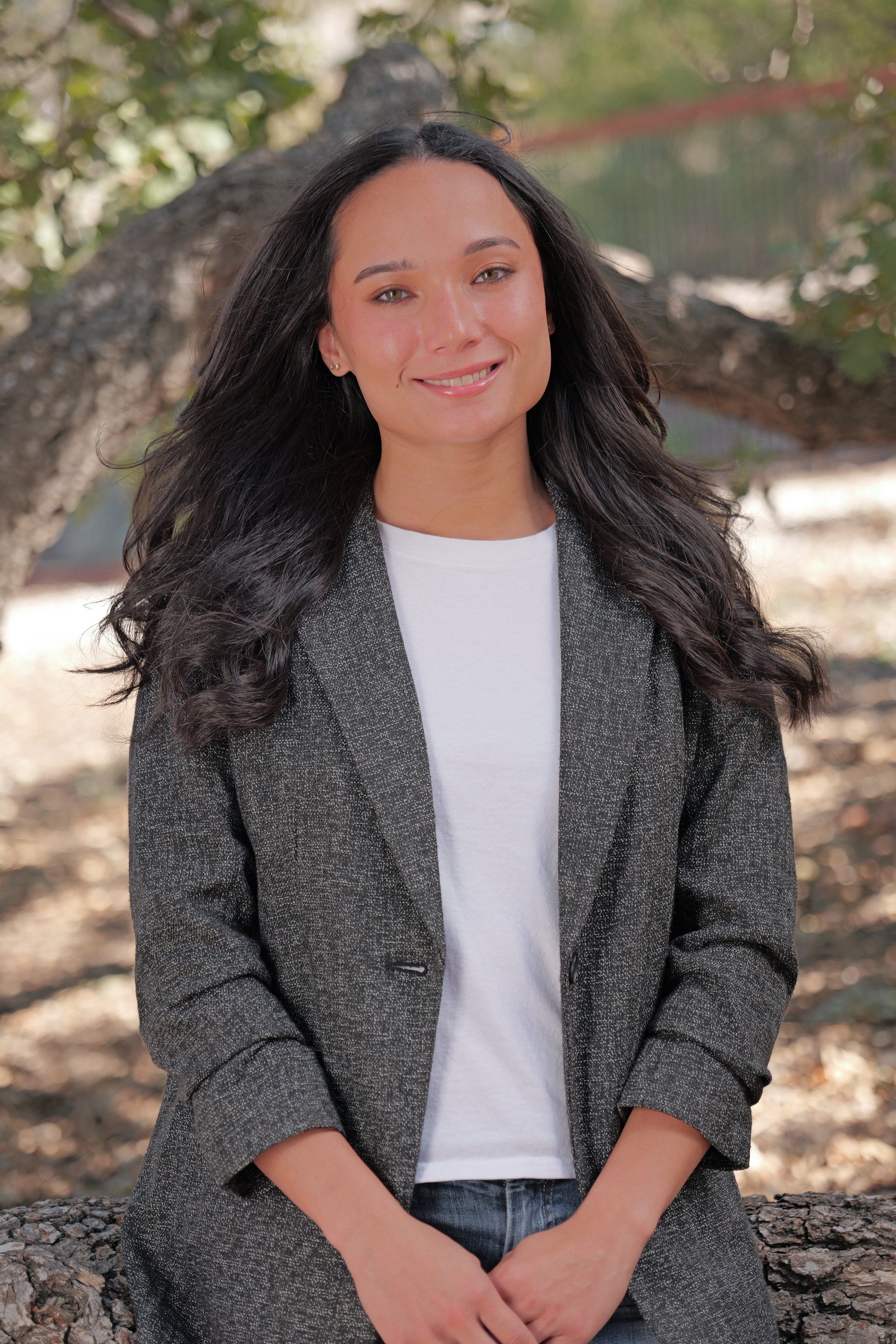 A young woman with dark hair, light skin, and blue eyes smiling outdoors, wearing a denim jacket and hoop earrings, with a blurred natural background.