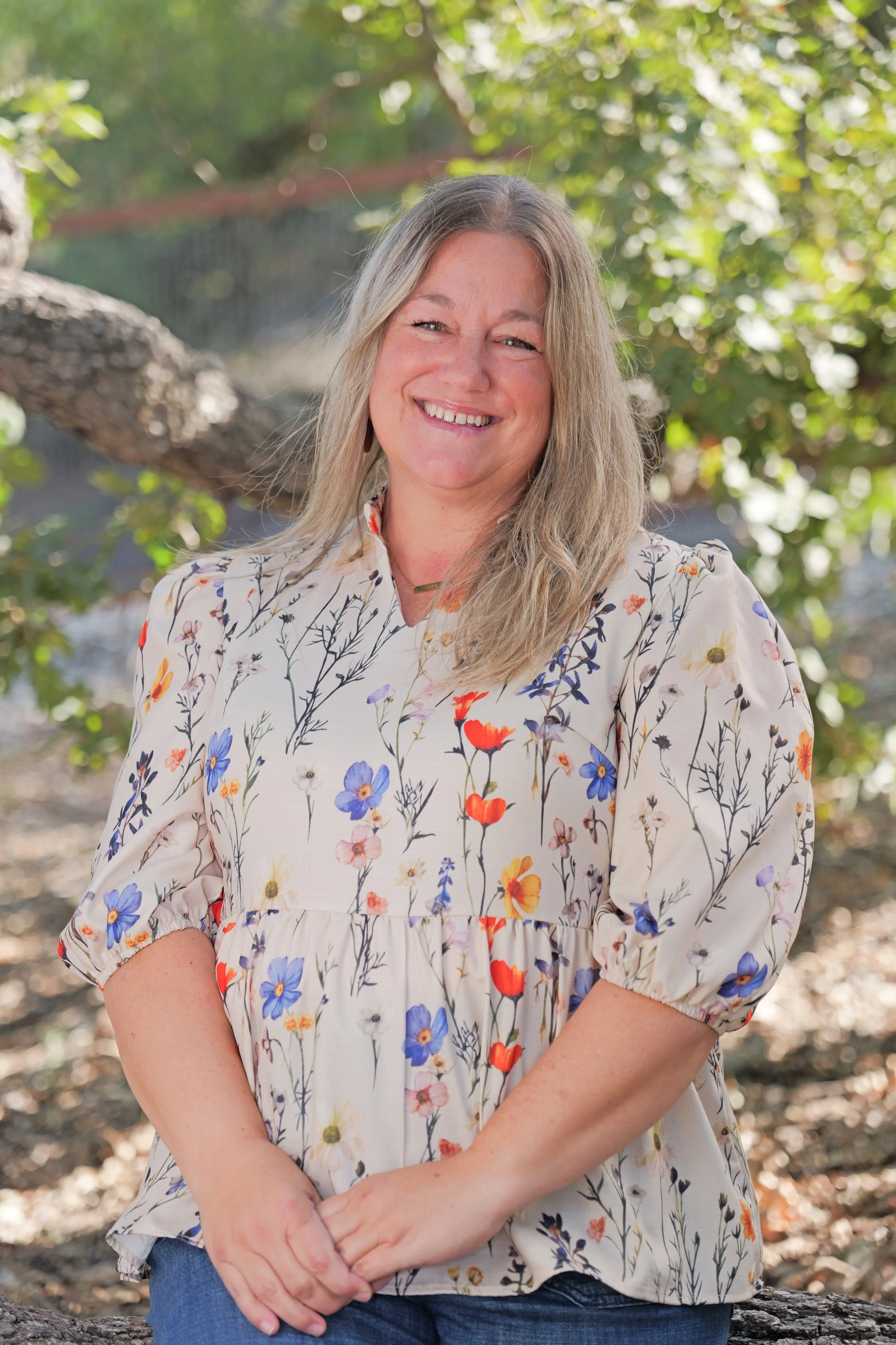 A smiling woman with blonde hair wearing a floral blouse and necklace, sitting outdoors with trees and sunlight in the background.