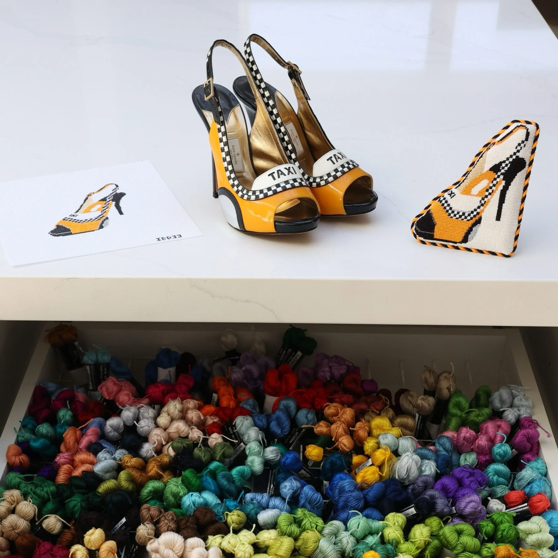 A display of a pair of yellow and black high-heeled shoes with a checkered pattern and an upper with 'TAXI' written on it, along with a matching finished needlepoint design, and the original hand painted canvas.