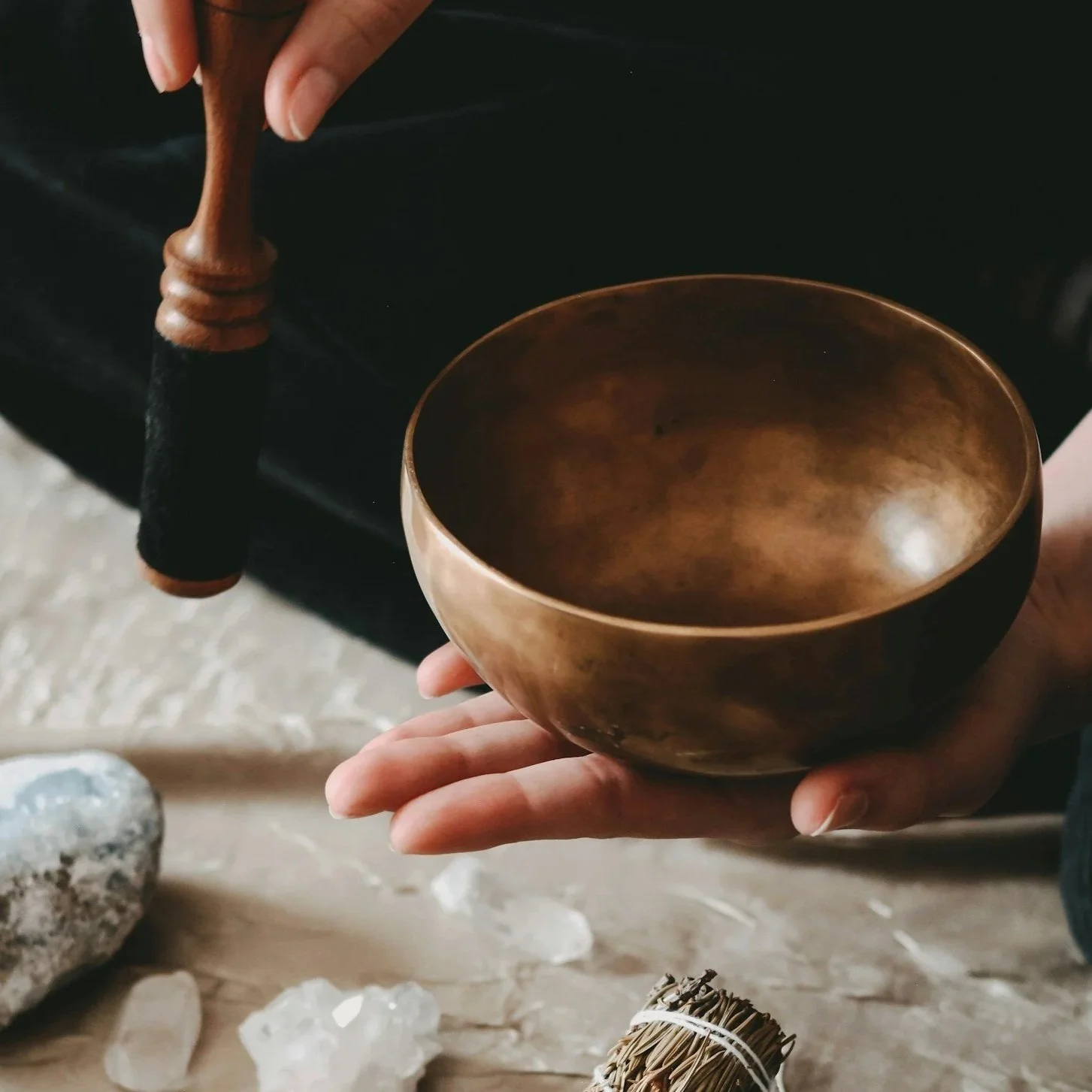 A hand holding a singing bowl with a wooden striker above it, with crystals and stones on a wooden surface below.