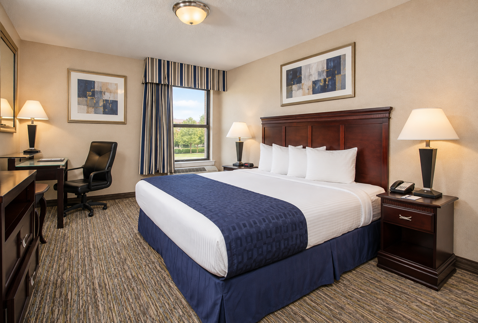 Hotel guest room with a king-size bed, navy bedding, wooden furniture, and natural window light.
