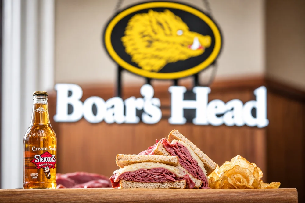 Deli sandwich with potato chips and a bottle of cream soda in front of a Boar’s Head sign.