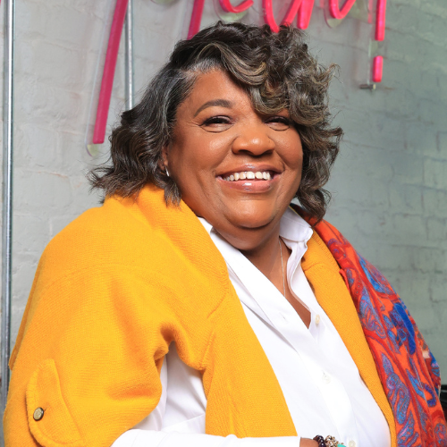 Smiling woman with curly gray hair wearing a yellow jacket and white shirt, standing in front of a brick wall with a neon sign.