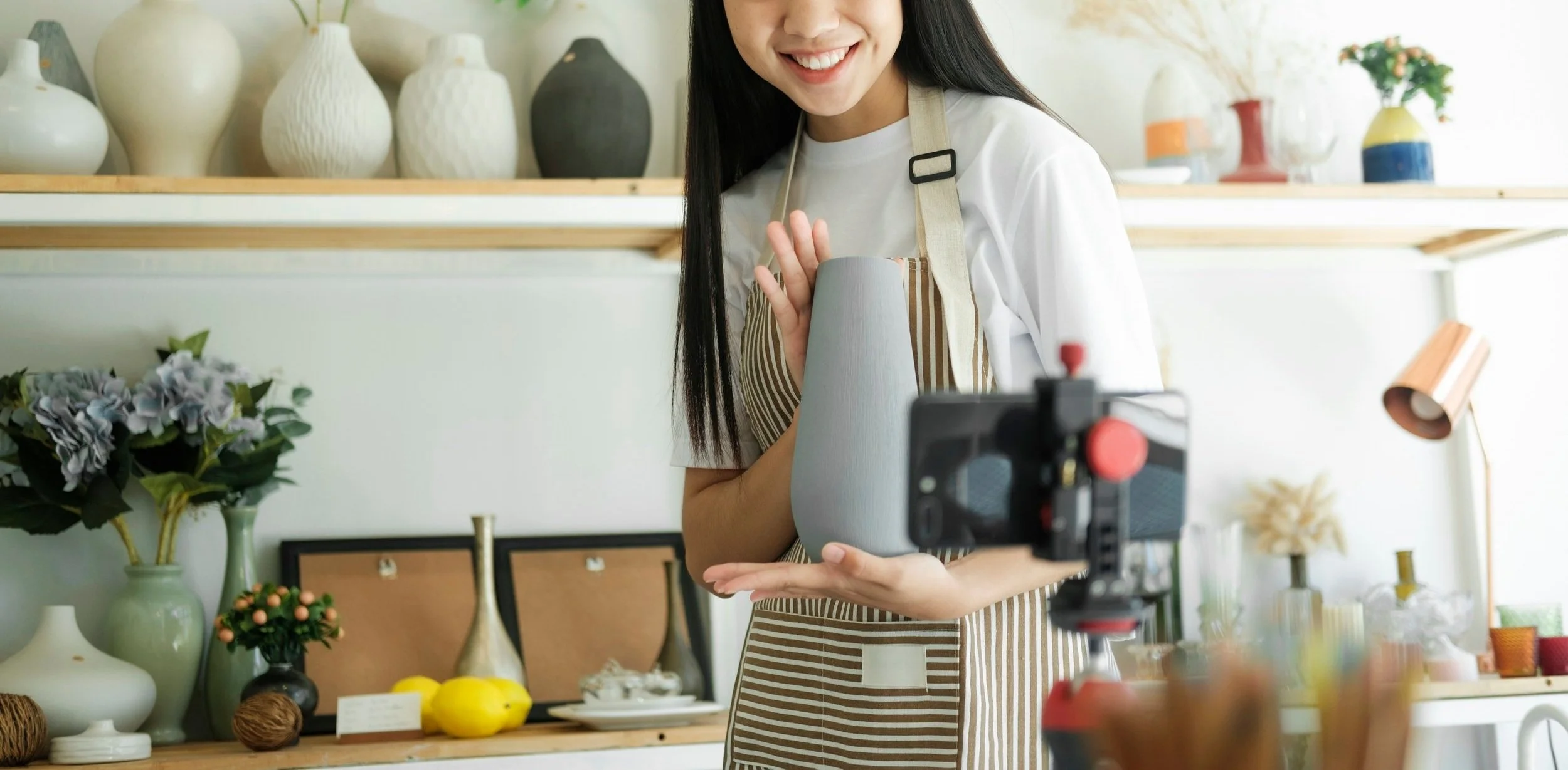 Woman in striped apron filming a ceramic vase with a camera in a decorated home studio.