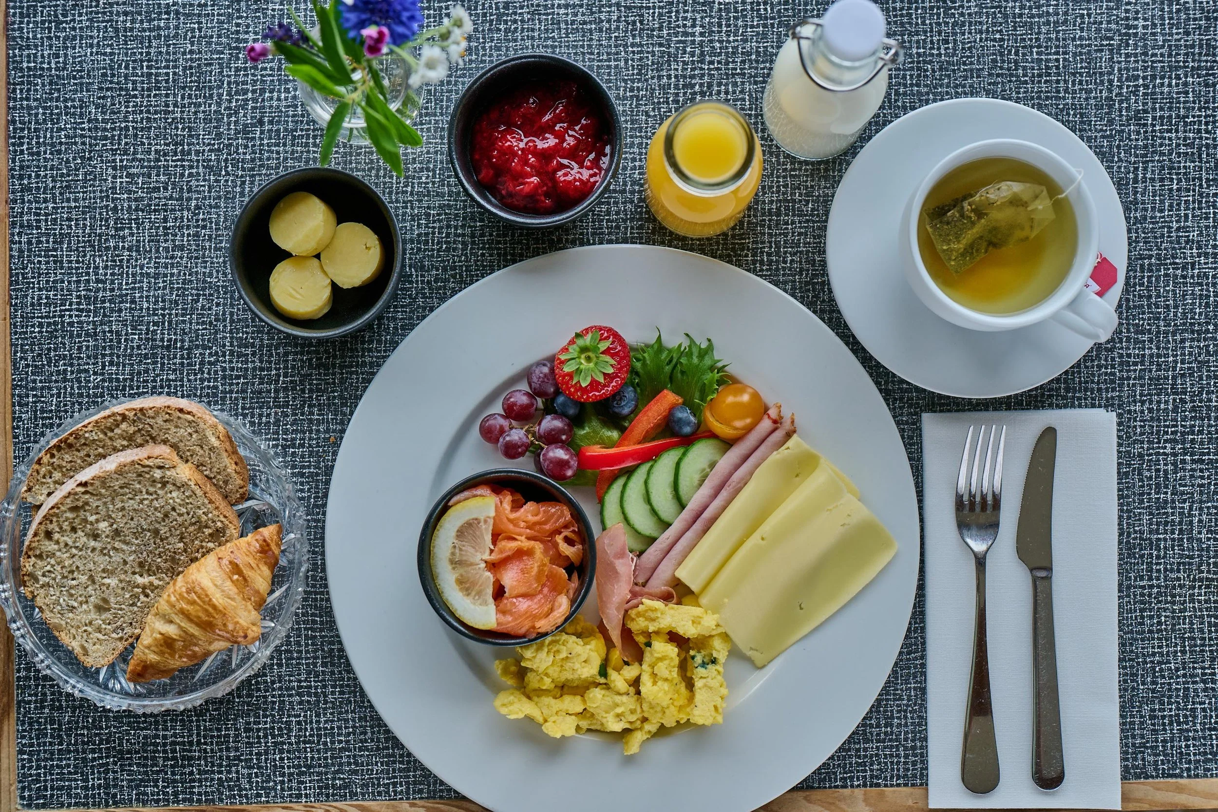 Frokost med skiver av brød, croissant, frukt, ost, pålegg, egg, juice og te på et duket bord.