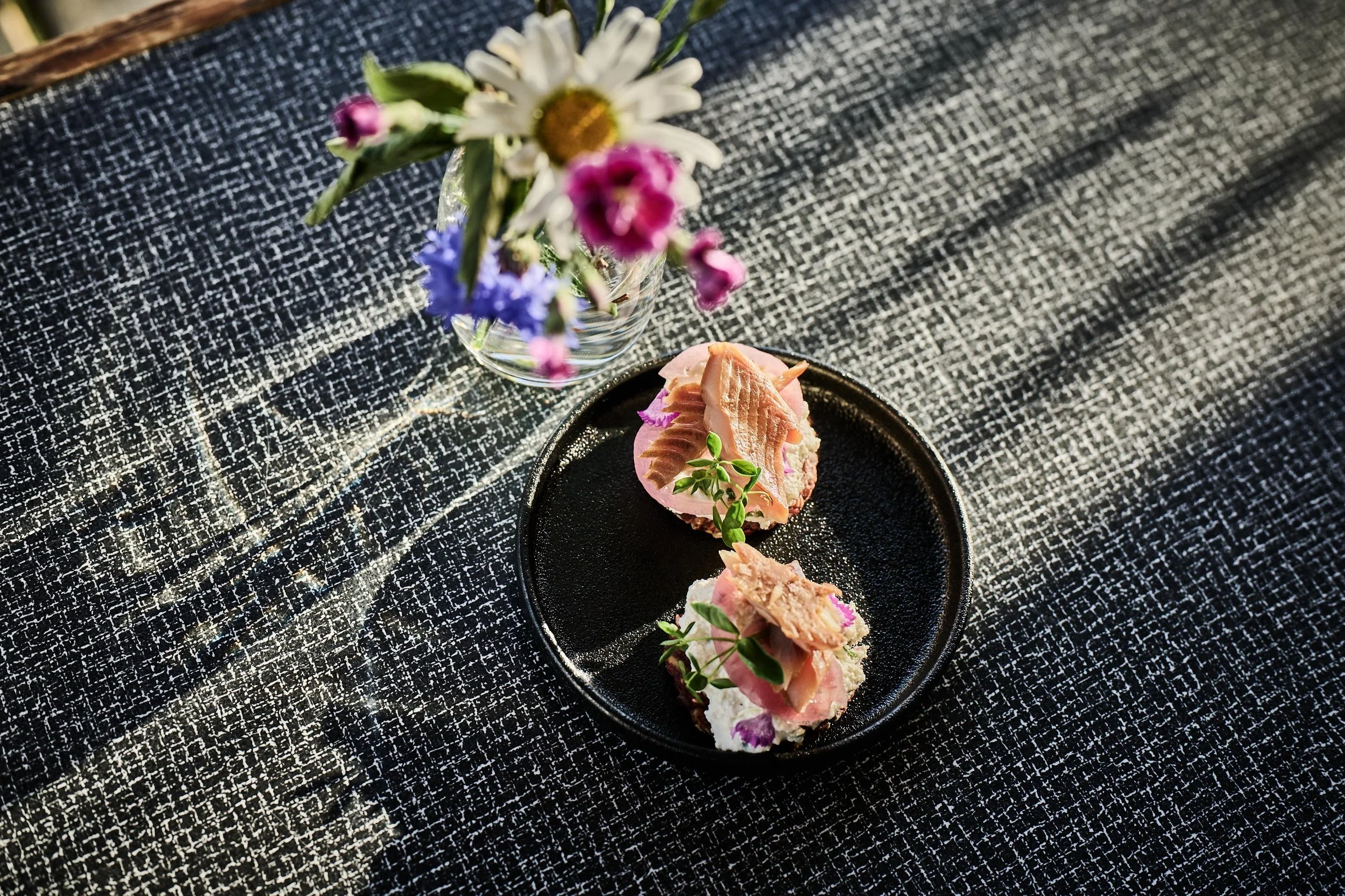 To smørbrød med tunfisk på en sort tallerken, pyntet med urter, plassert på en svart og hvit vevet duk på et bord, med en vase med blomster i bakgrunnen.
