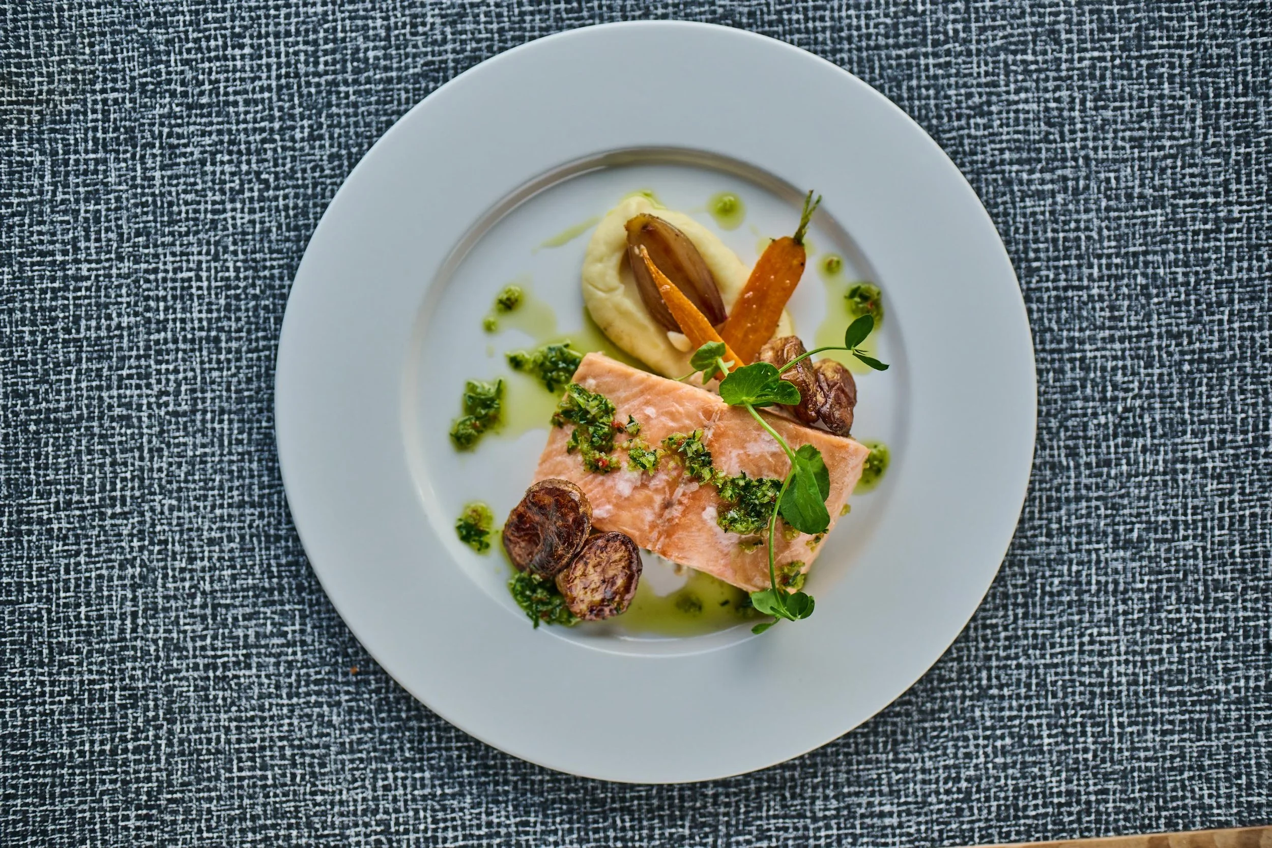 Porsjon med rosa laks, potetstappe, sopp og grønne urter på en hvit tallerken på en gråtekstilet bordduk.