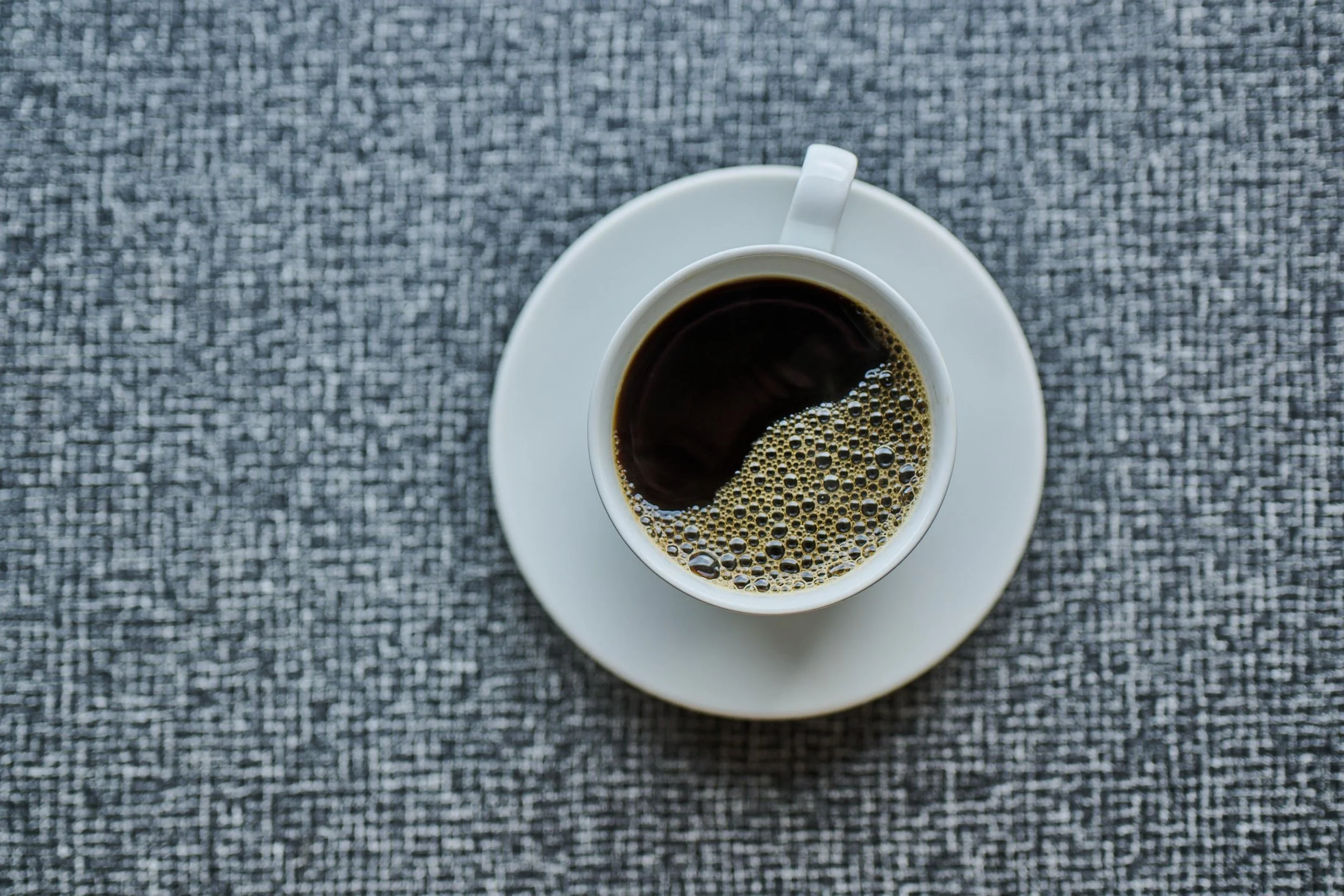 En kopp med svart kaffe på et hvitt fat, sett ovenfra, plassert på en grå teksturert overflate.