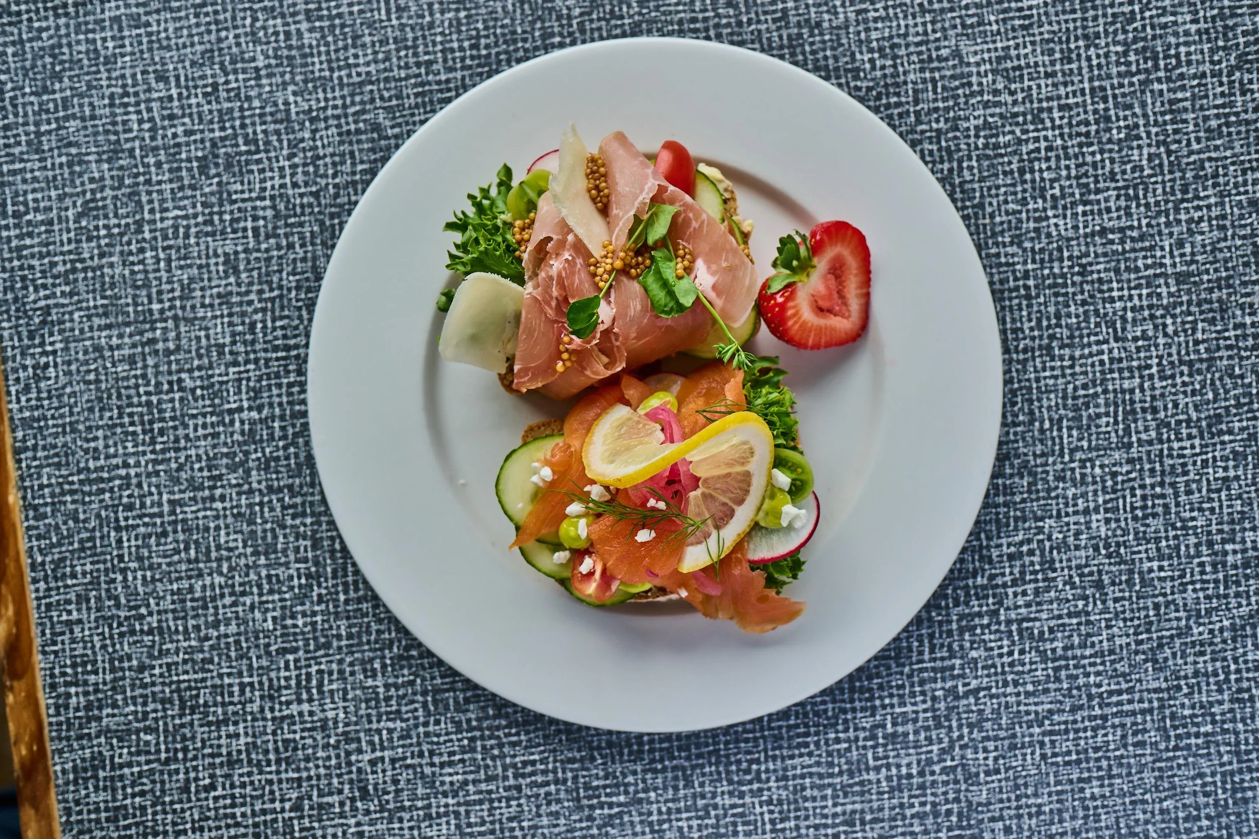 En tallerken med salat, spekeskinke, og skivede grønnsaker, med jordbær på siden, plassert på en hvit tallerken på en sort og hvit teksturert duk.