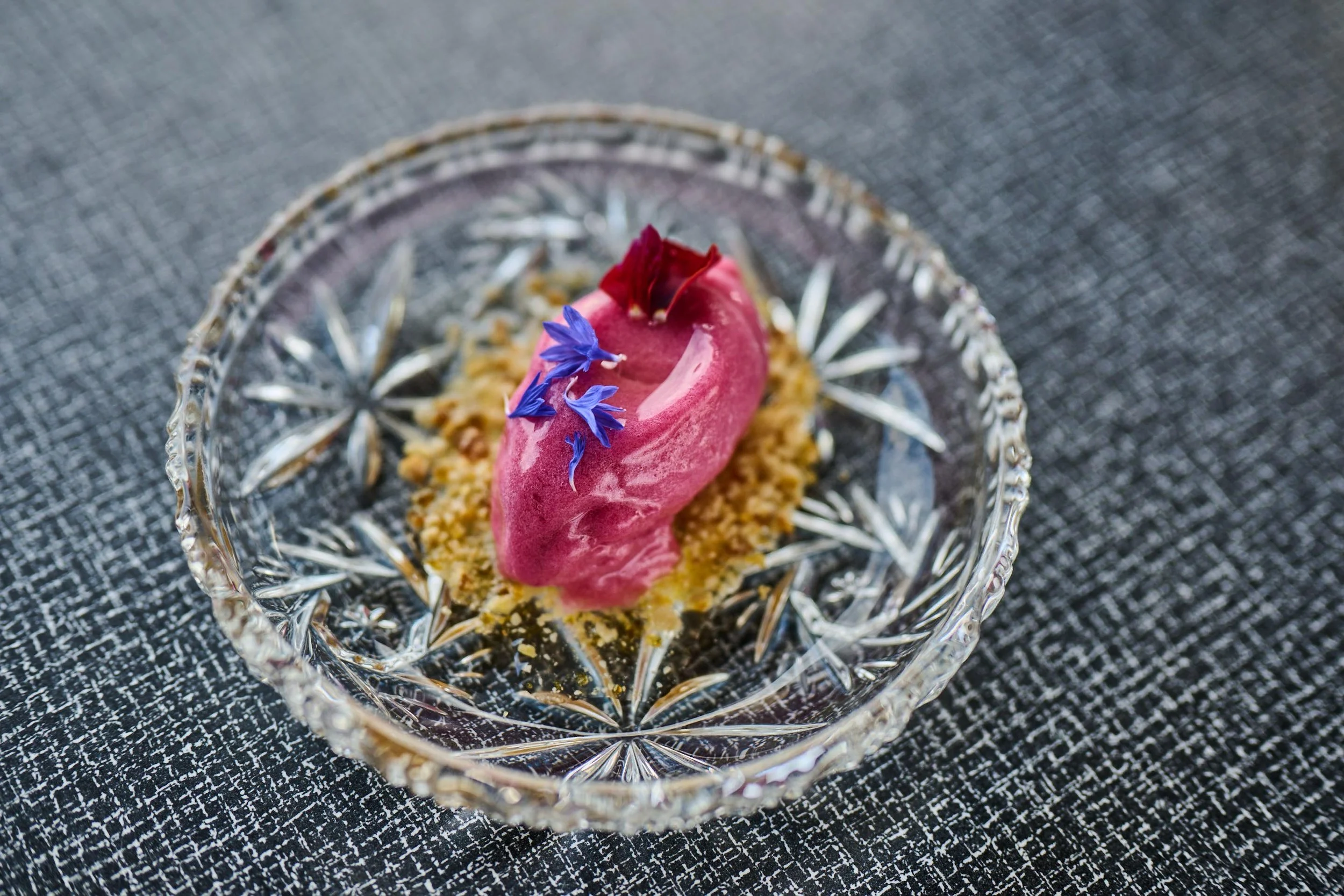 En glasskrem med rosa farge, pyntet med blå og rød blomster, på en glassfat med krystallmønster.