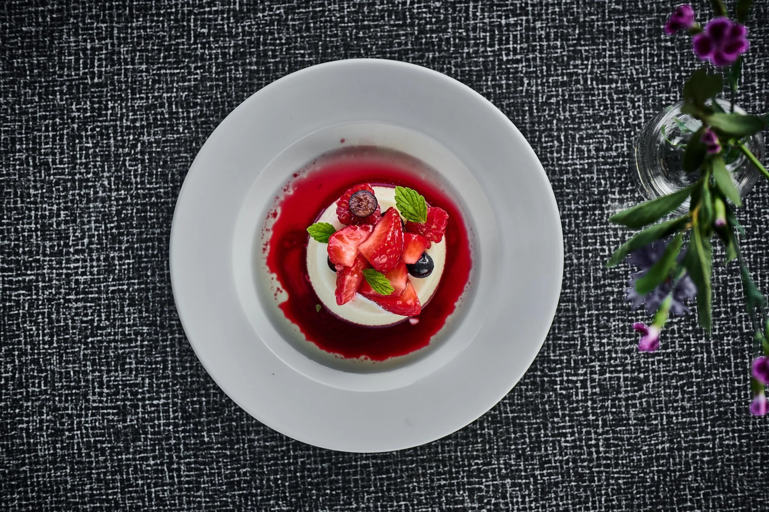 Kremet dessert med frukt og grønne blader, omgitt av rød saus på en hvit tallerken.