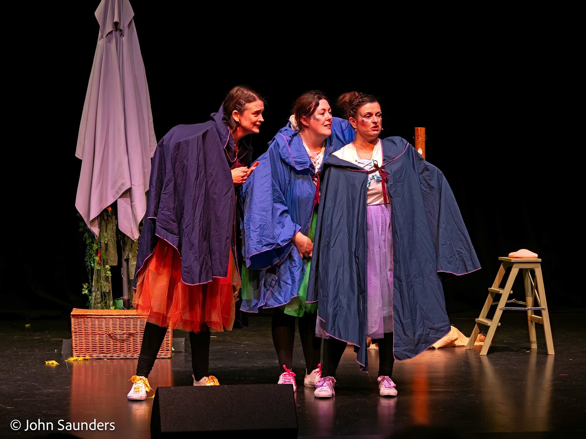 Four women in colorful raincoats performing on stage with humorous expressions, surrounded by stage props including a wicker basket, a stool with a hat, and a large umbrella.