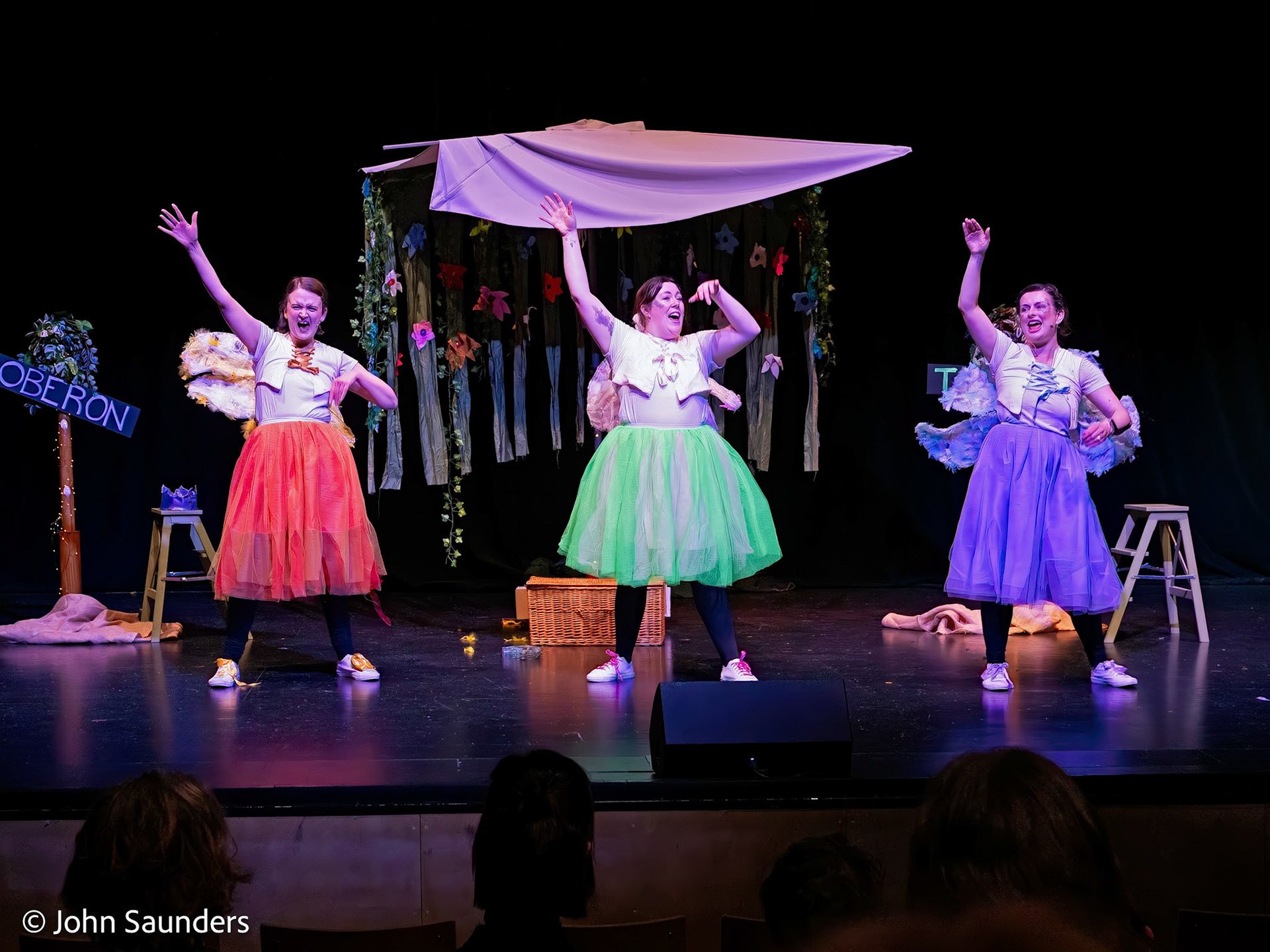 Three women performing on stage dressed in colorful skirts and white tops, with a backdrop of flowers and trees, during a theatrical production.