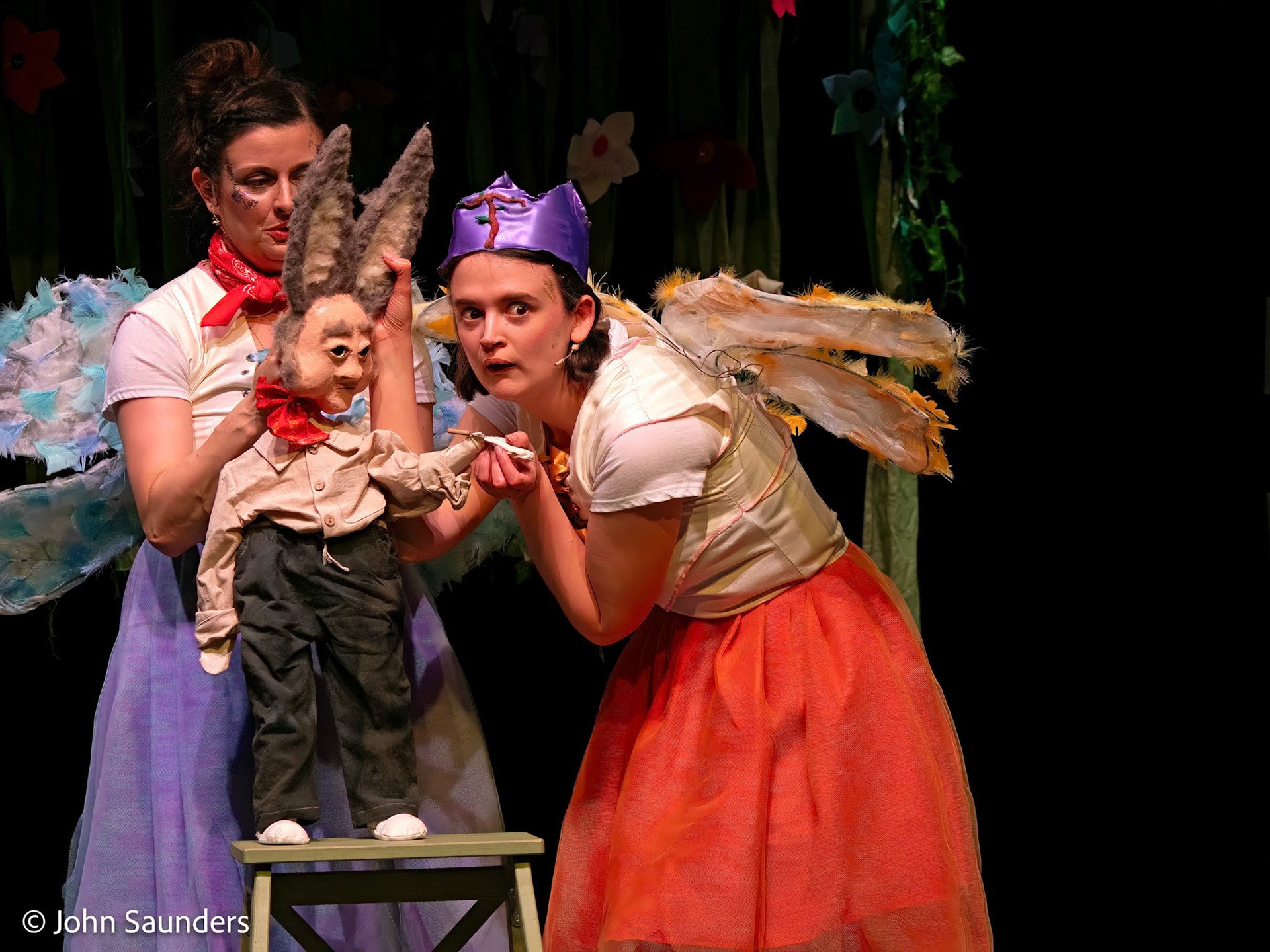 Two women in colorful costumes with wings, and a puppet with donkey ears standing on a small platform, on stage during a theatrical performance.