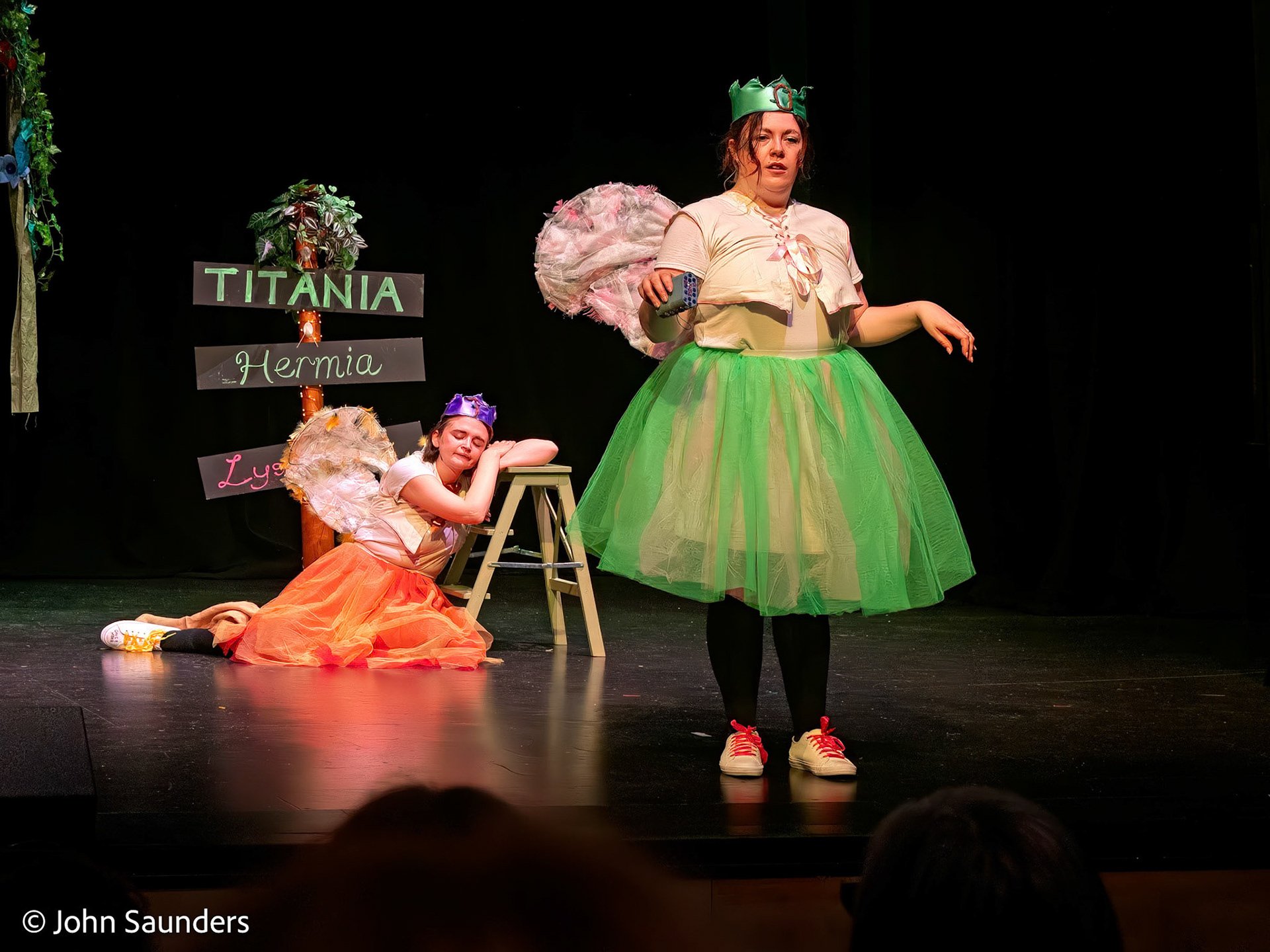 Stage play scene with three actresses in fairy princess costumes, one standing in green tutu and two sitting in blond wigs and fairy wings, with a sign in the background reading "TITANIA, Hermia, Lysander" and a black curtain.