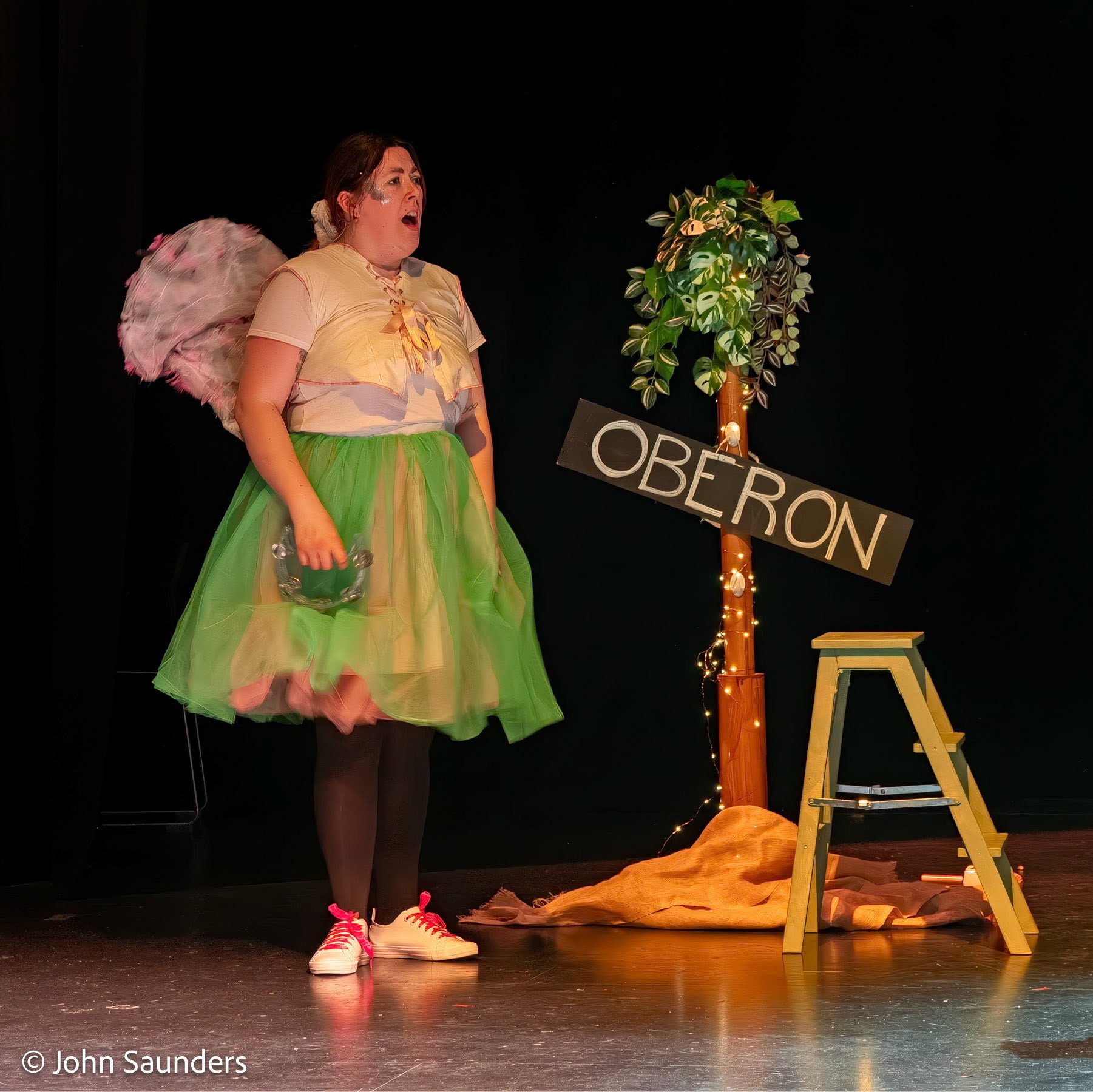 A woman in a colorful costume standing on stage with a surprised expression, holding a tambourine, next to a sign that says "OBÉRON" and a decorative tree with lights.