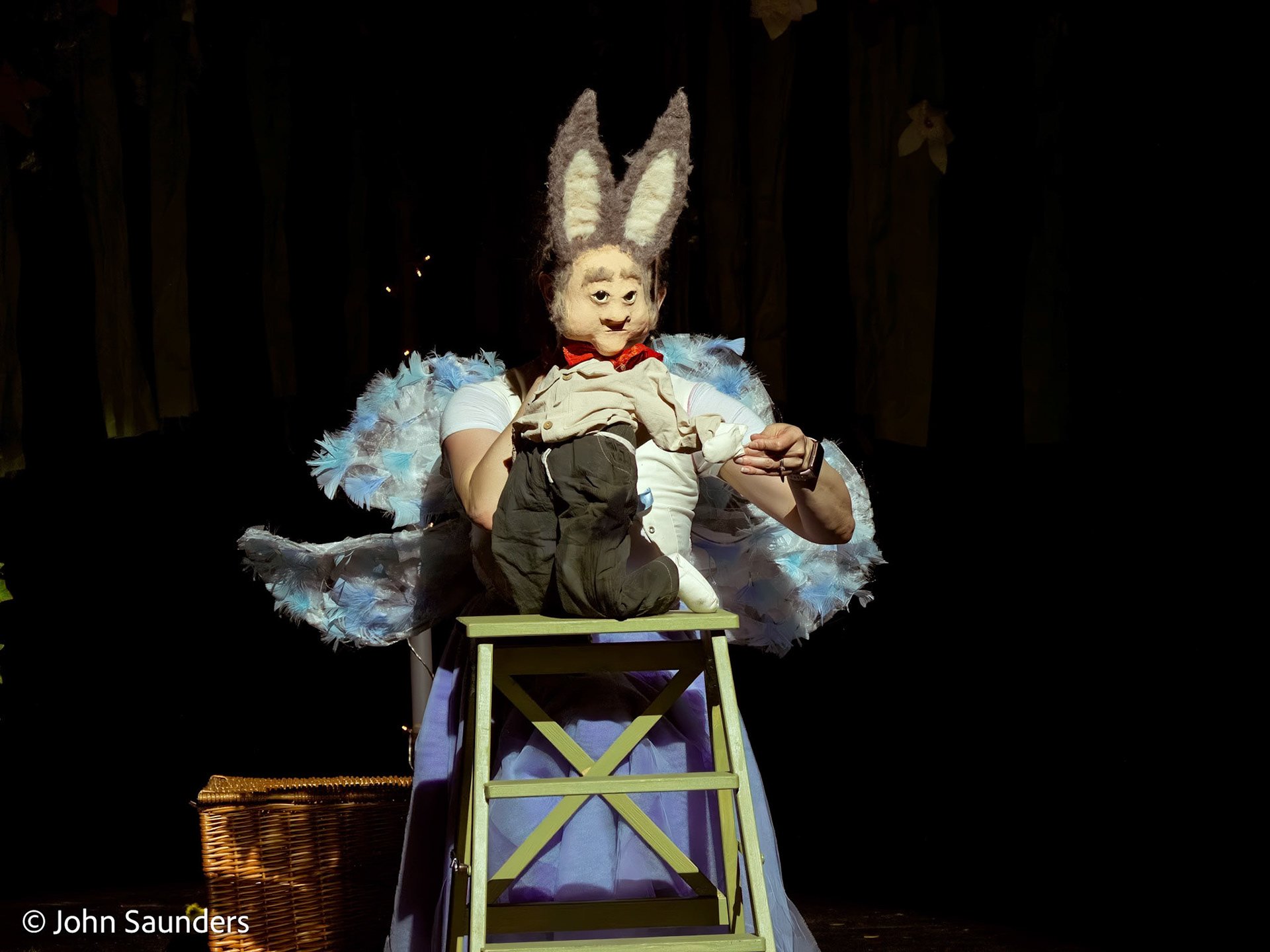 A puppet character with large donkey ears, wearing a red bowtie and a beige shirt, is sitting on a small green stool with a puppet stage background. The puppet's clothes include dark pants and white shoes, and it has a painted smiling face.