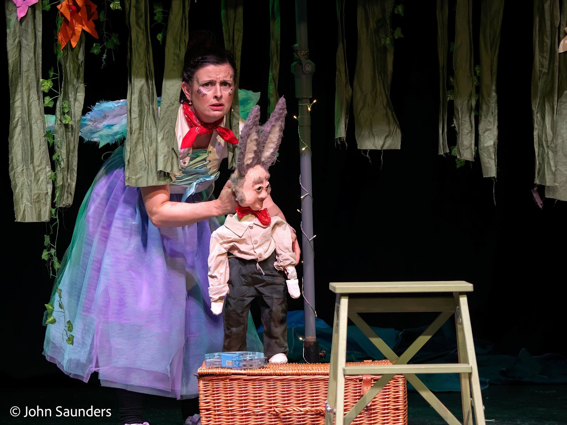 A woman dressed in a costume with wings and makeup appears distressed while holding a puppet of a donkey in a theatrical stage setting with forest backdrop.