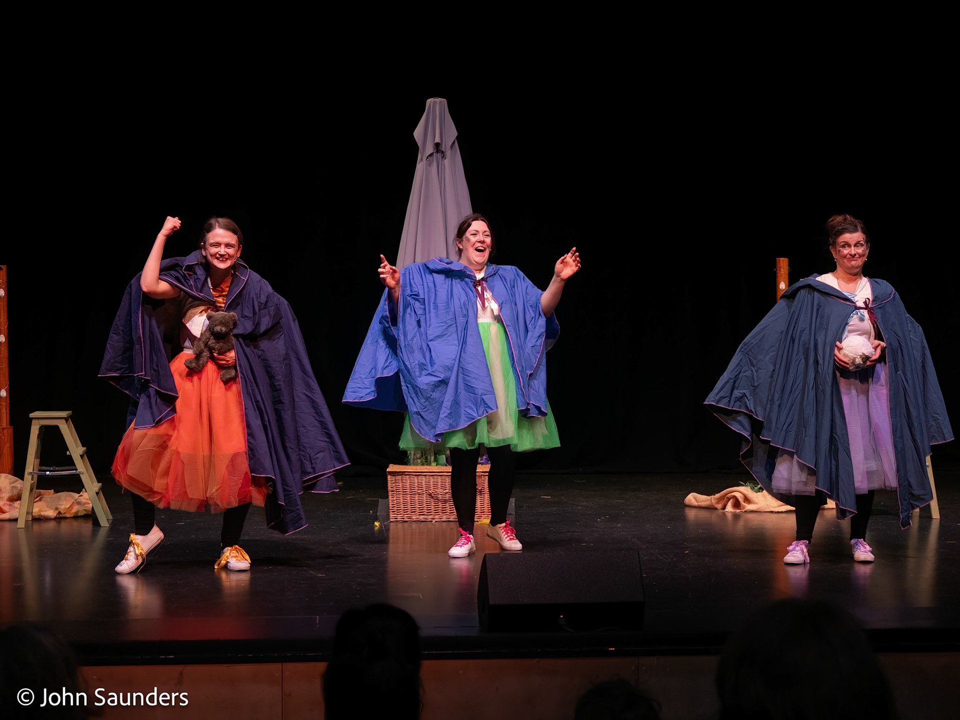 Three women performing on stage, wearing colorful dresses and capes, with props and set pieces including a picnic basket, chairs, and a large object resembling a ball or rock.