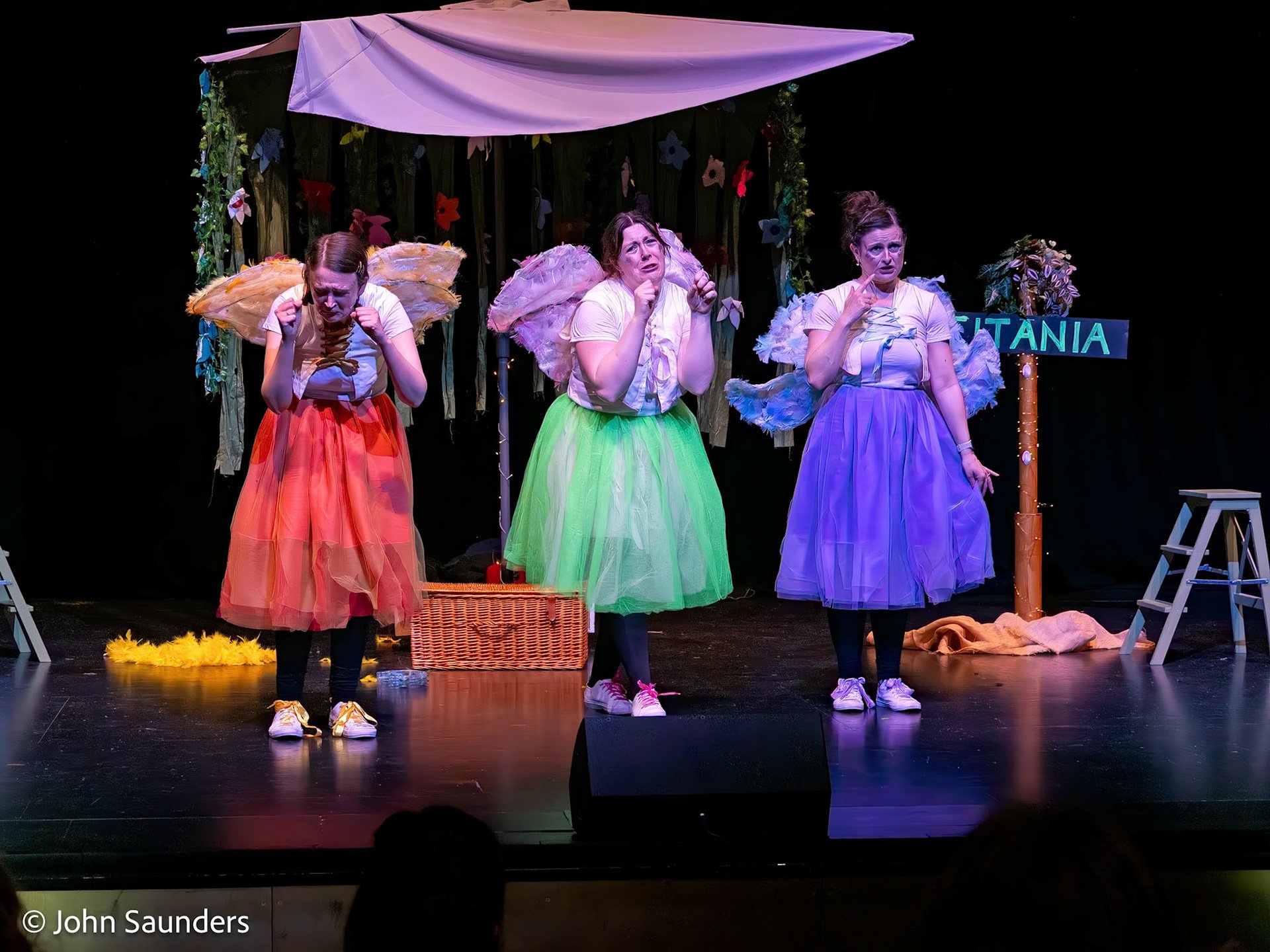 Three women dressed as fairies with colorful skirts and wings perform on stage in a theatrical play. They are standing in front of a decorated backdrop with flowers and a sign that reads 'TITANIA'.