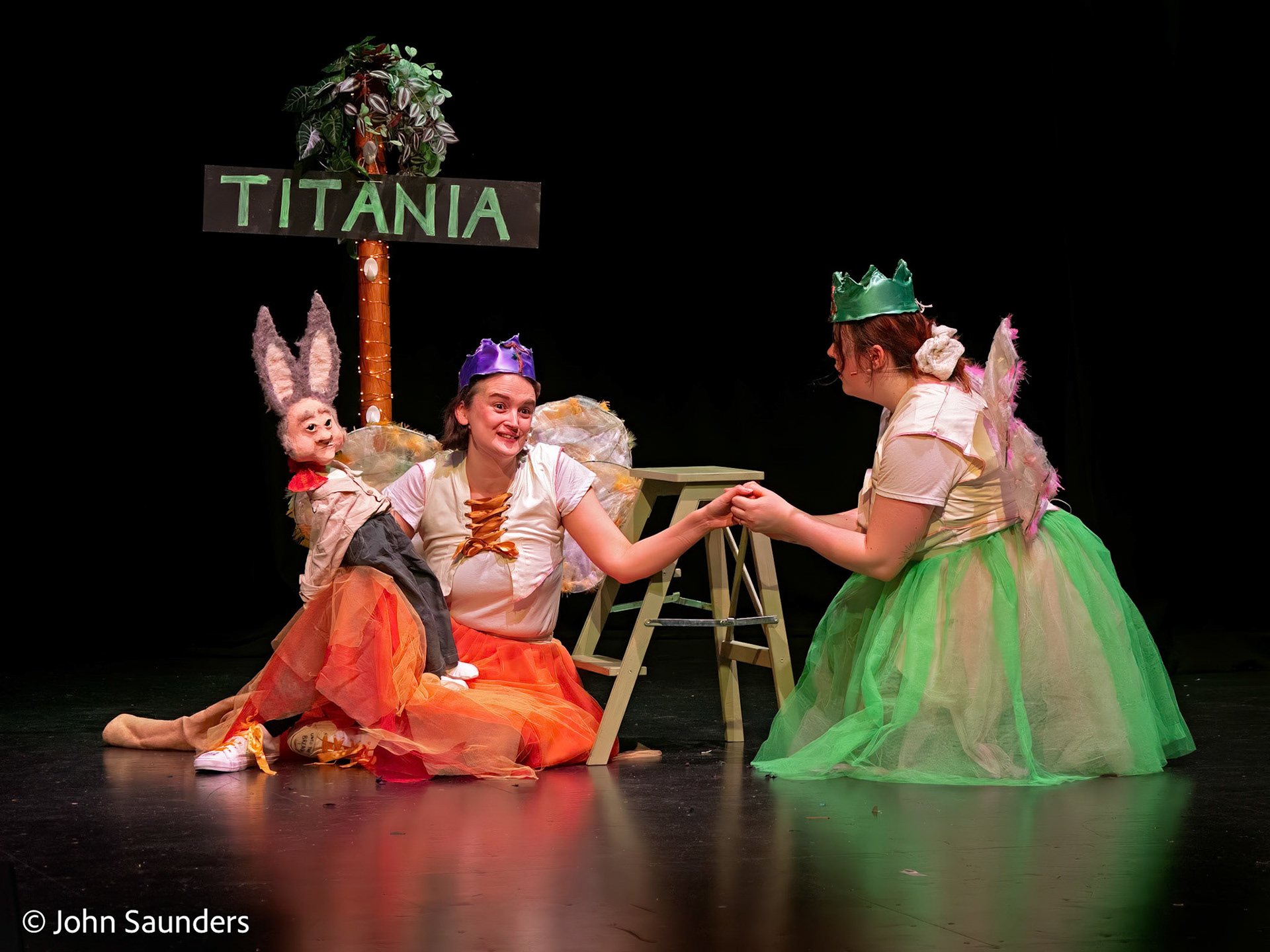 Two women in colorful fantasy costumes with wings and crown, one kneeling and the other standing, on stage holding hands, with a sign reading "Titânia" and a puppet donkey, during a theatrical performance.