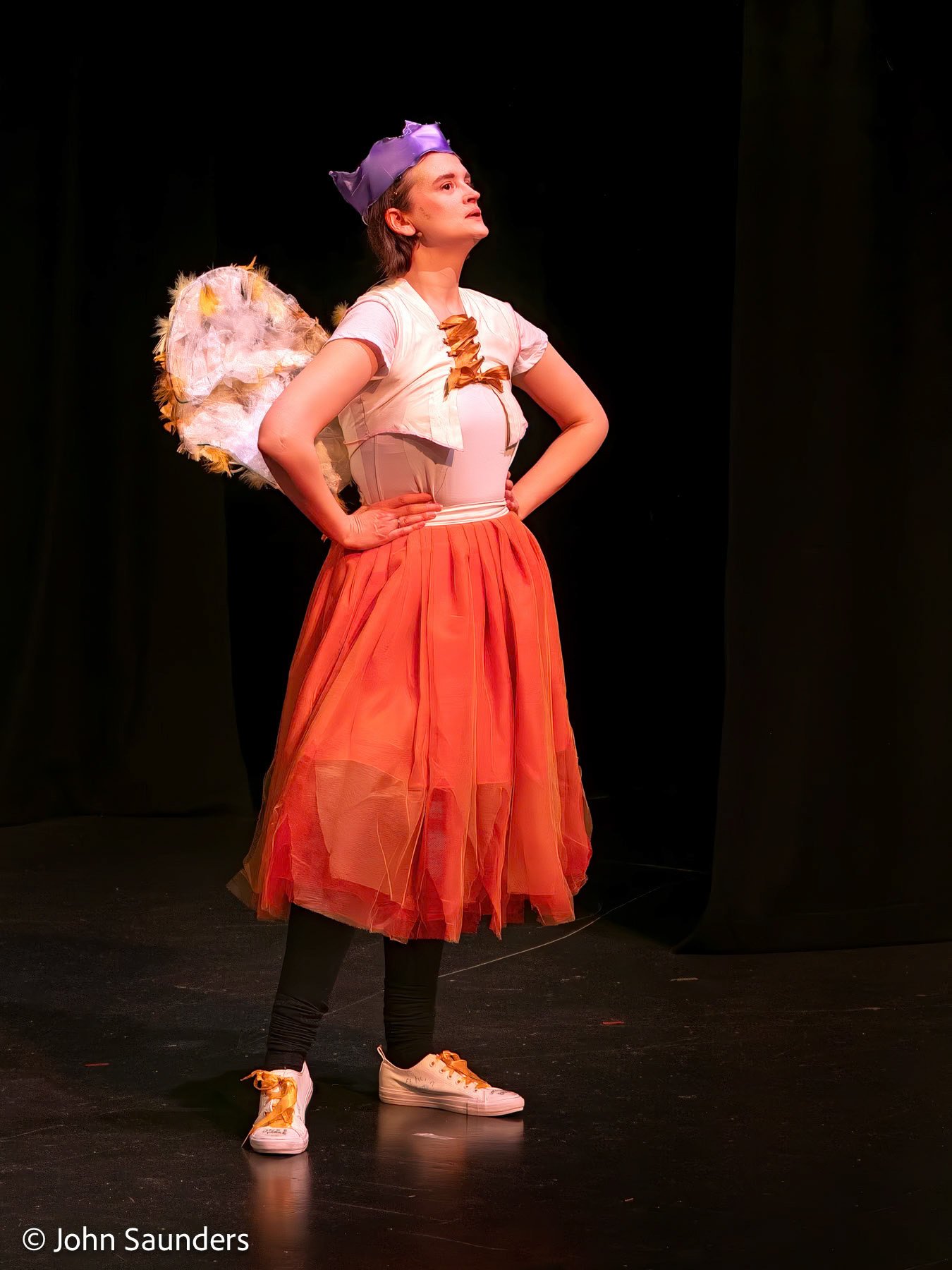 Person dressed as a fairy on stage with hands on hips, wearing a purple crown, white top, orange tulle skirt, angel wings, black leggings, and white sneakers.