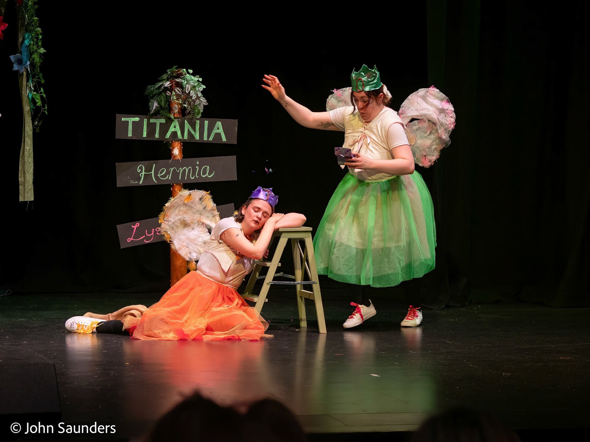 Two actors dressed as fairies performing on stage, one lying on the floor with a purple crown and orange skirt, the other standing with a green skirt, crown, and fairy wings, with a sign reading "TITANIA Hermia Lysander" in the background.