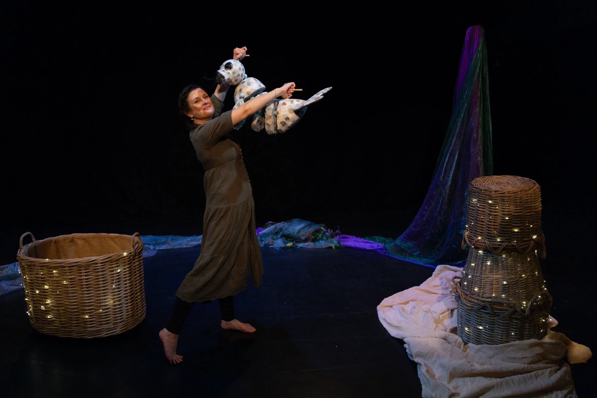Woman performing a play on stage with arts and crafts decorations, including wicker baskets and fabric, against a black background.