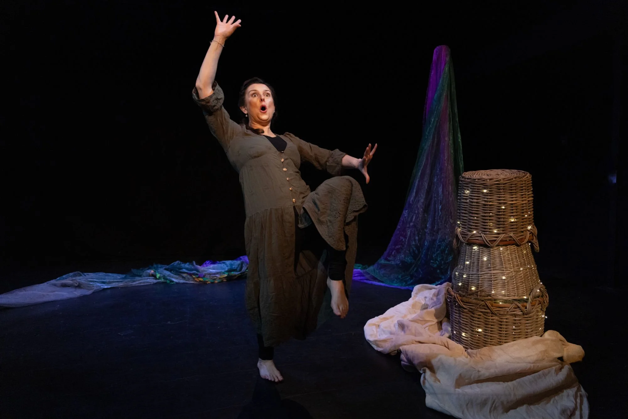 A woman with an expressive face and arm raised performing on a stage decorated with colorful fabric, wicker basket with string lights, and scattered cloths.