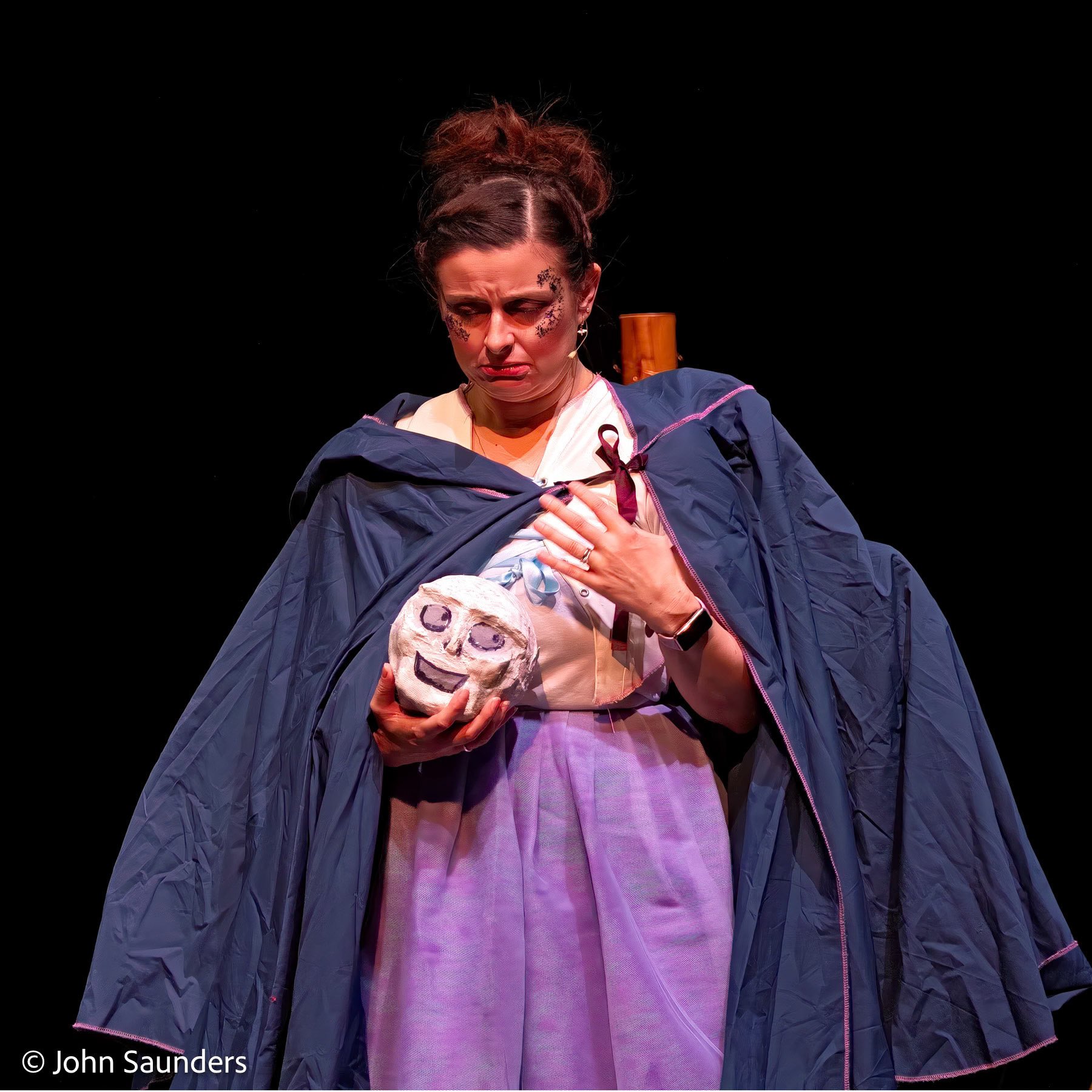 A woman dressed in a costume holds a painted face mask and appears emotional, standing against a dark background.