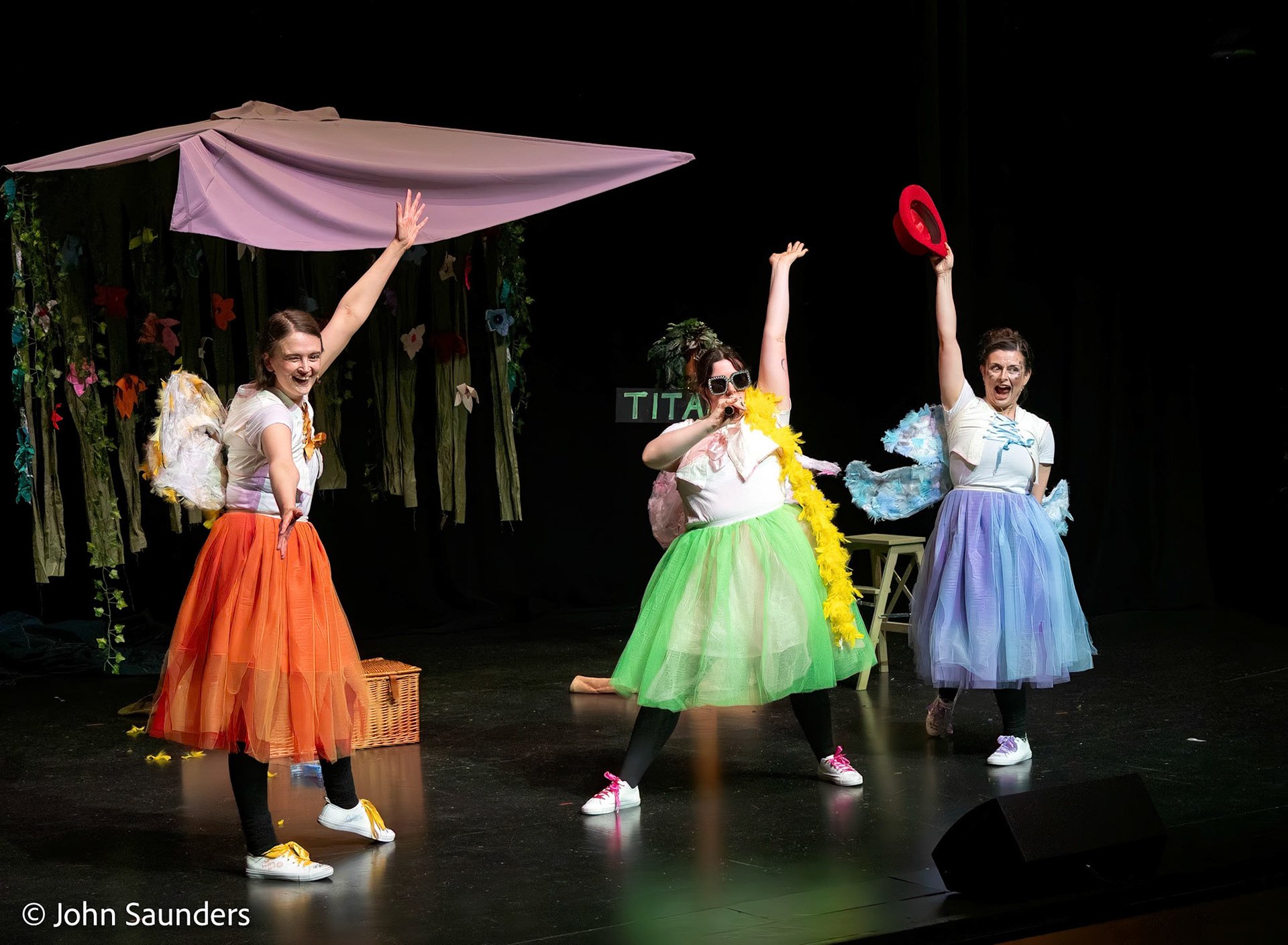 Three women dressed in colorful costumes performing on stage with a backdrop of flowers and a large umbrella prop.