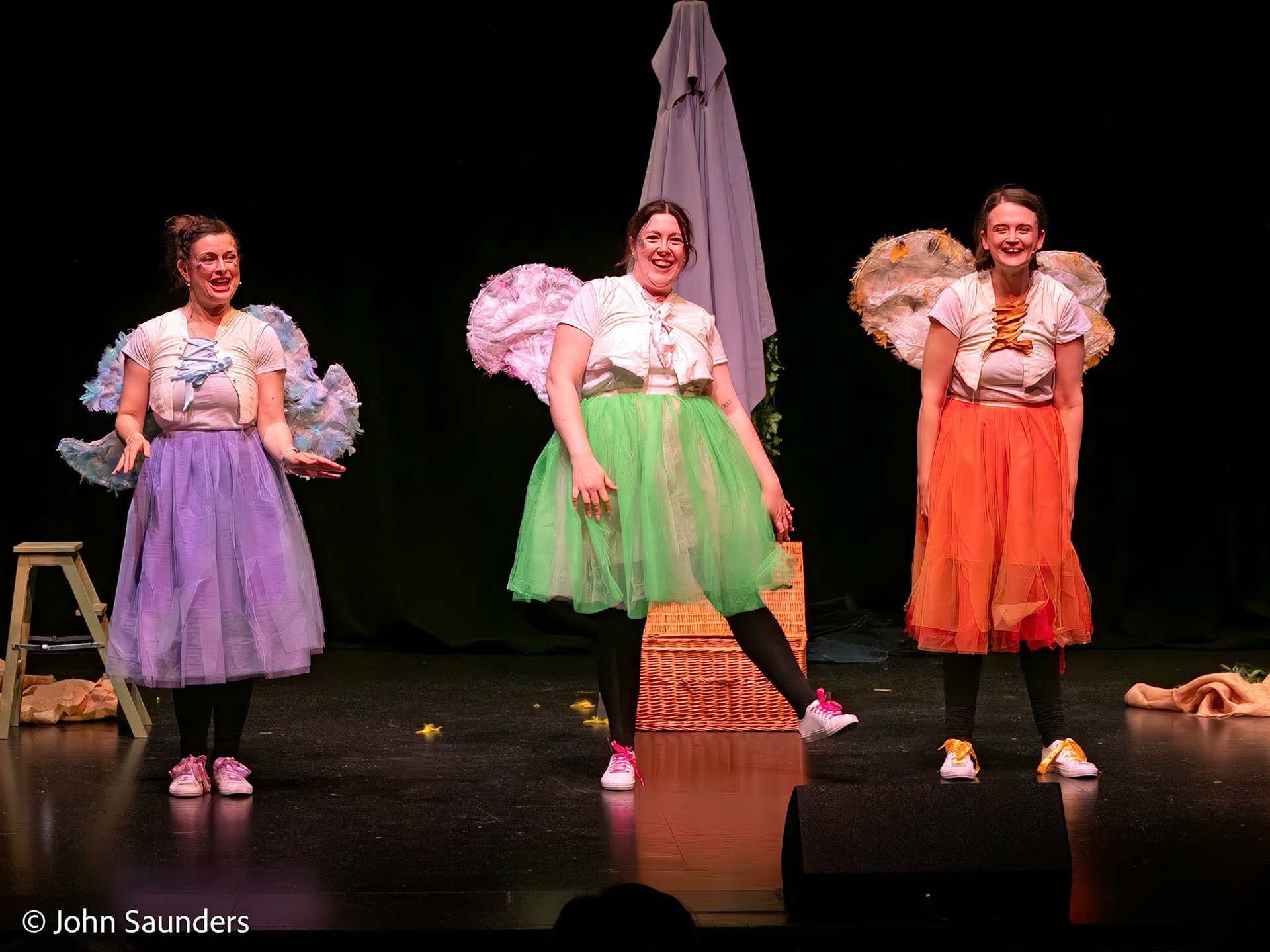 Three women on stage dressed as colorful fairies with wings, wearing pastel and bright skirts, performing in a play or dance. They are smiling and standing in front of a black background with stage props.