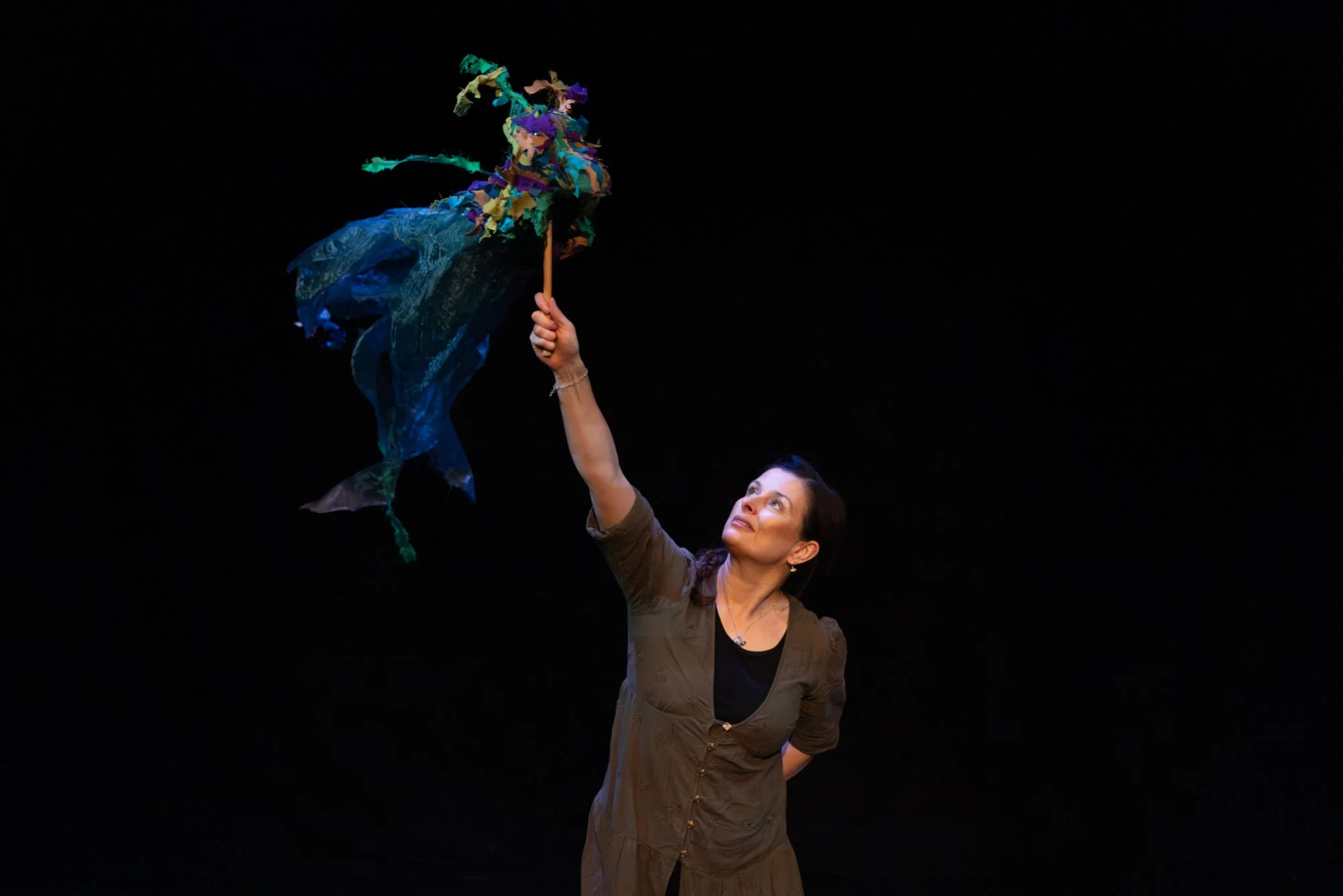 A woman performing with a colorful, flowing ribbon Kelpie water horse puppet on a black stage.