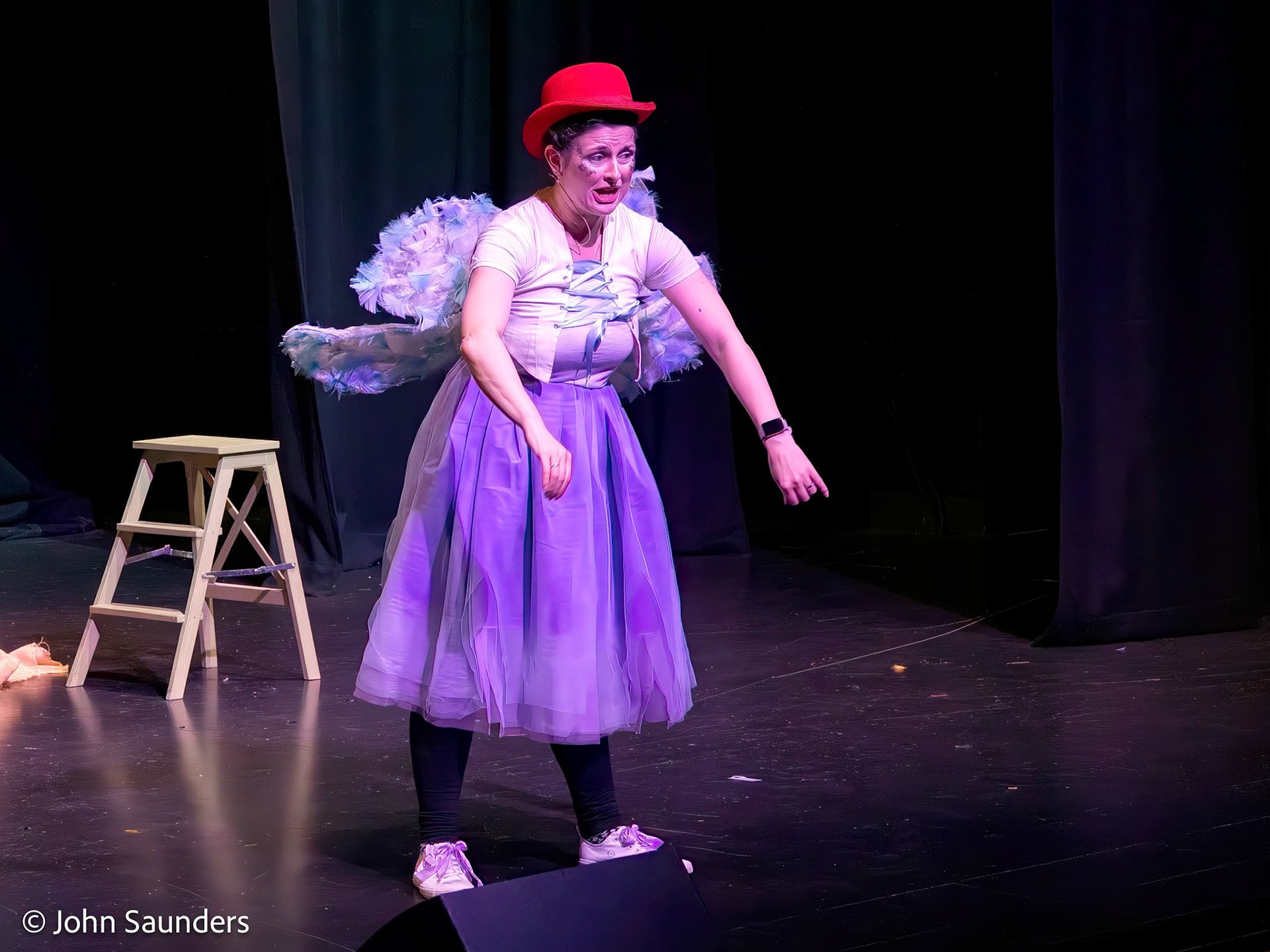 A performer on stage dressed in a colorful costume, wearing a red hat, purple skirt, and white top with wings, expressing a dramatic or expressive moment.