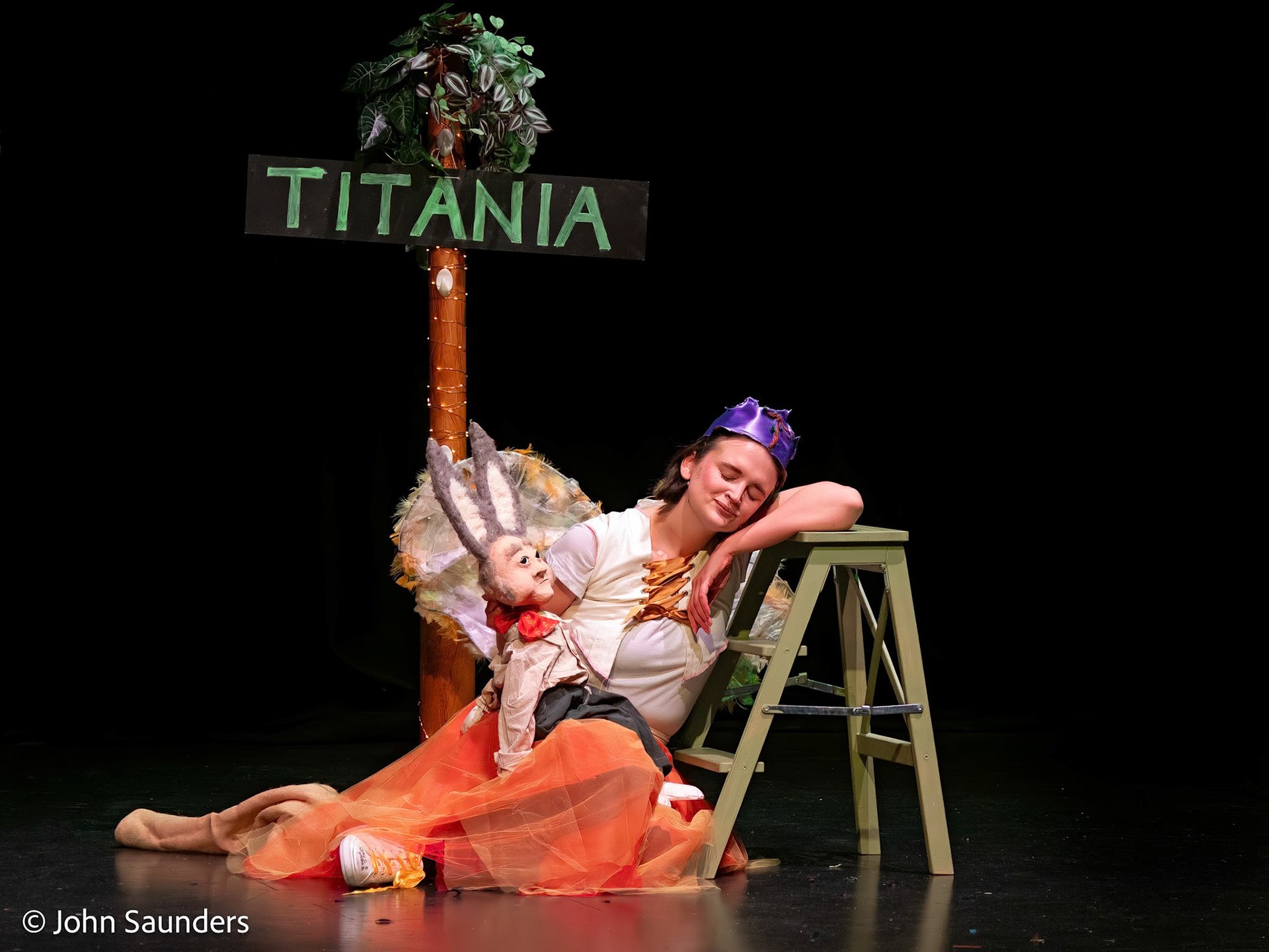 A young woman dressed as a fairy sleeping with her head resting on a small table on stage, with a puppet donkey in front of her, and a sign reading 'TITANIA' in the background. The stage is dark with focused lighting.