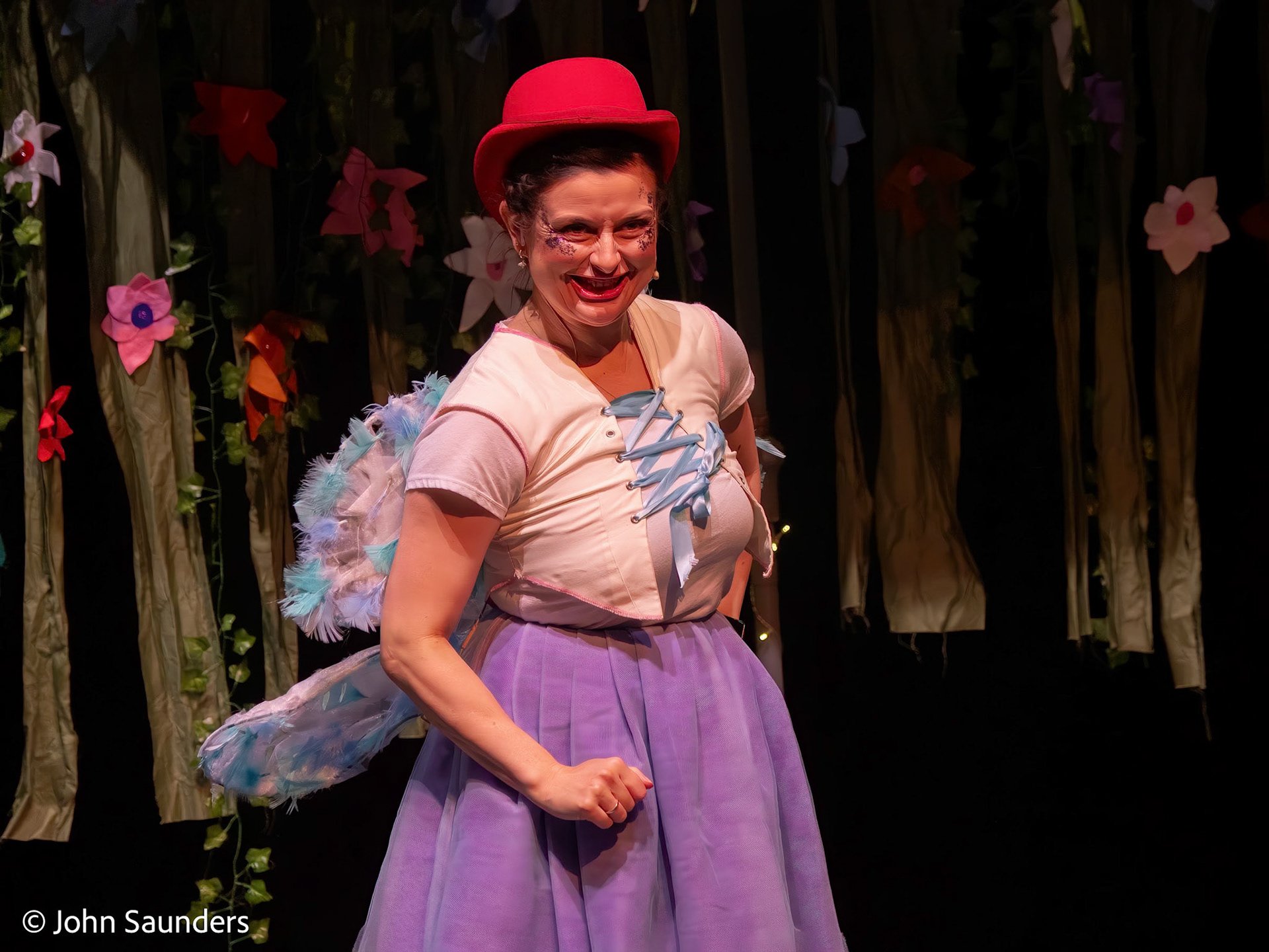 A woman in a colorful costume standing on stage, smiling with a forest-themed backdrop with flowers.