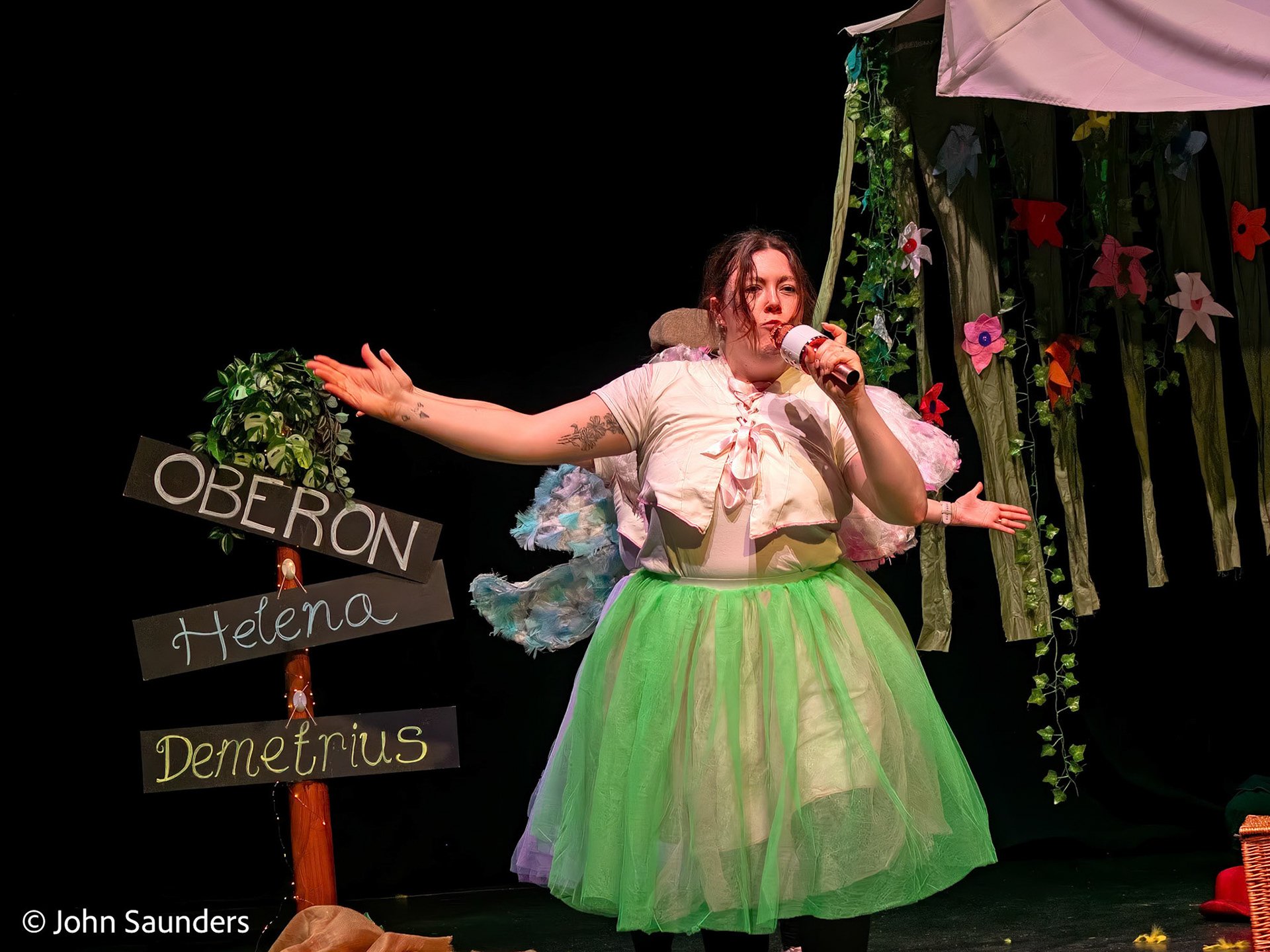 A woman dressed as a fairy standing on stage with a green tulle skirt, holding a microphone, with a sign that reads 'Oberon Helena Demetrius' and a decorated background with flowers and foliage.