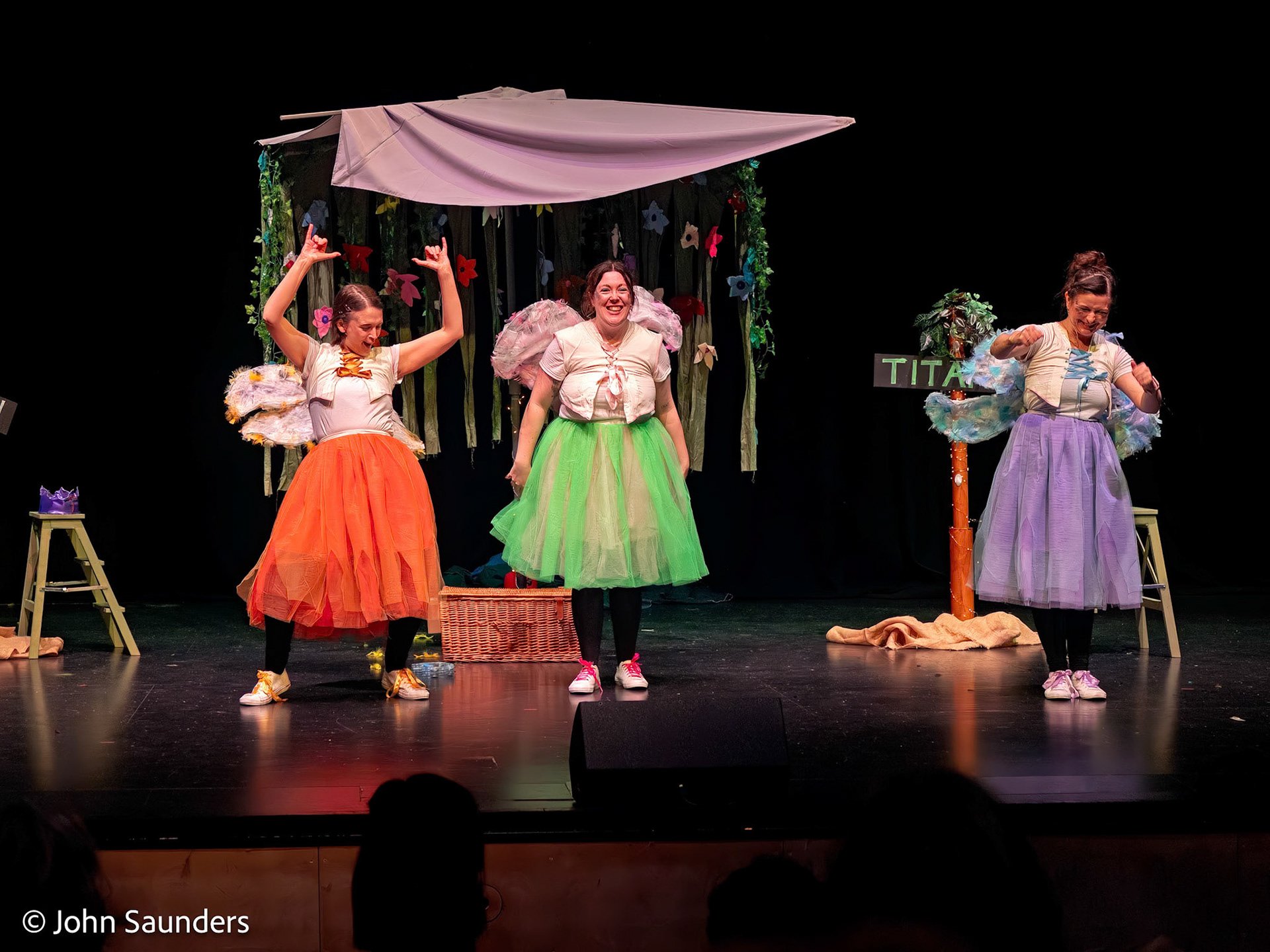 Three women performing on a stage in colorful costumes, with a flower garden set and a black backdrop.