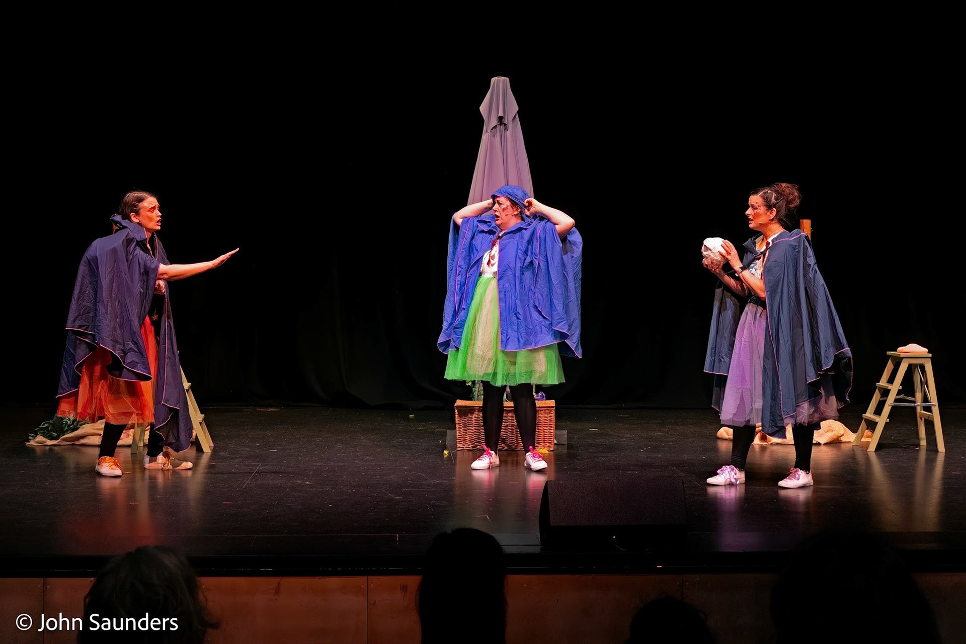 Three women performing a theatrical play on stage, dressed in colorful costumes with robes, in front of a black backdrop, with stage props including baskets, a stool, and a small table.
