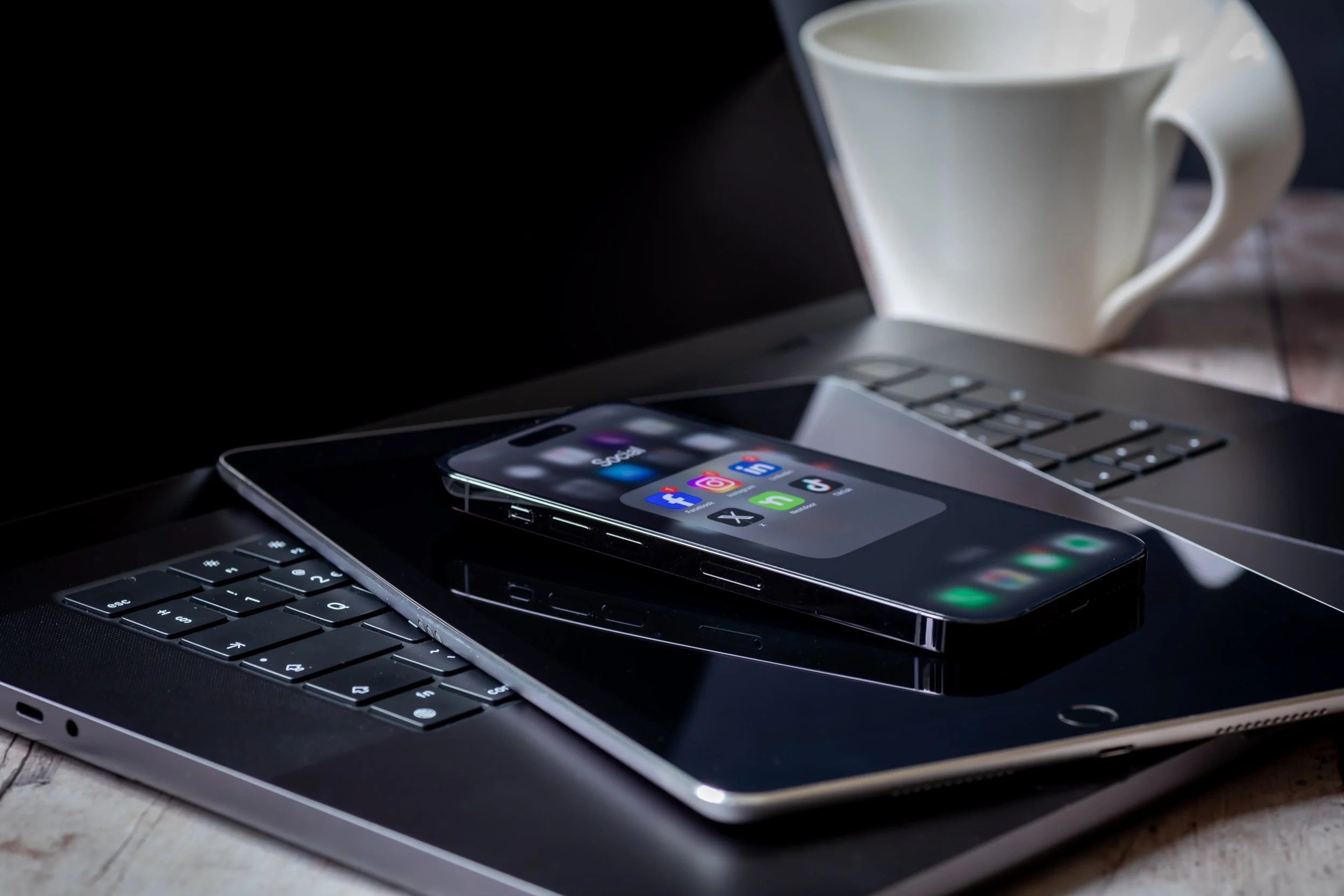 A smartphone, laptop, and tablet stacked on a wooden surface with a white mug in the background.