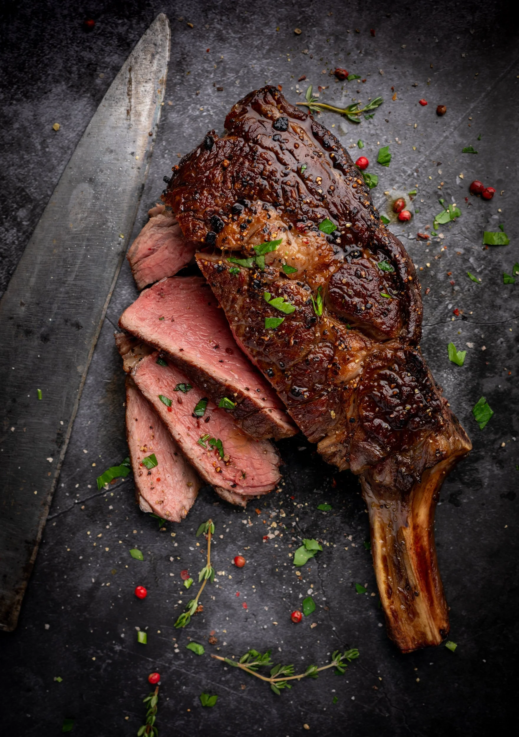 Fine art photograph of sliced cote de boeuf scattered with herbs and pink peppercorns with French carving knife at The Rising Sun Milland