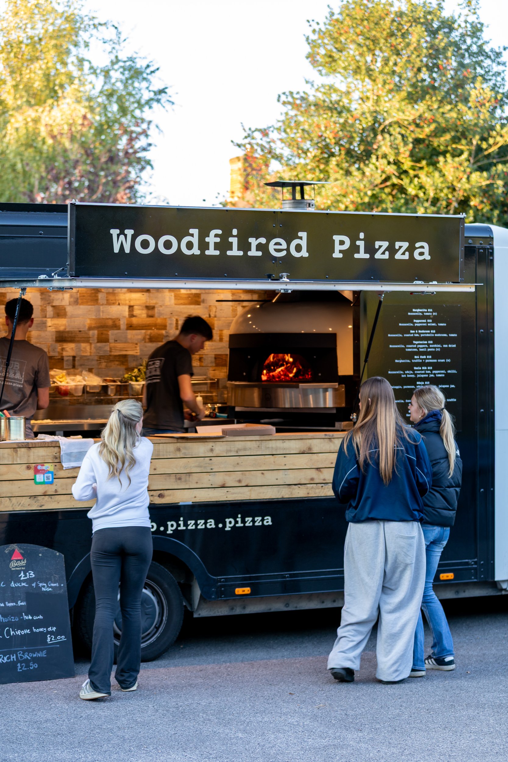 Three girls choosing their pizzas at the Club Pizza van parked at The Crossways Inn, Churt, Surrey. Lifestyle & food photography by Sequoia Studios
