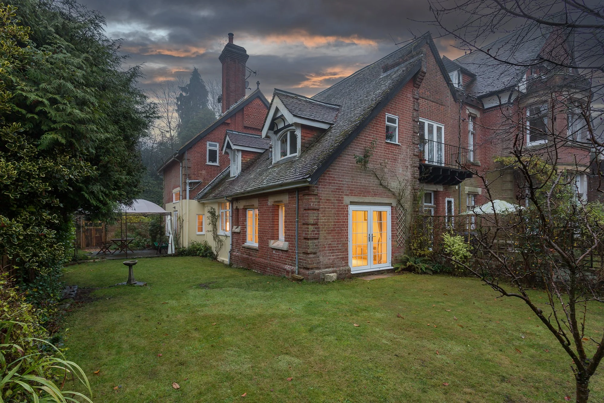 A brick house with lit windows and a door, surrounded by a green lawn and trees, at dusk with cloudy sky.