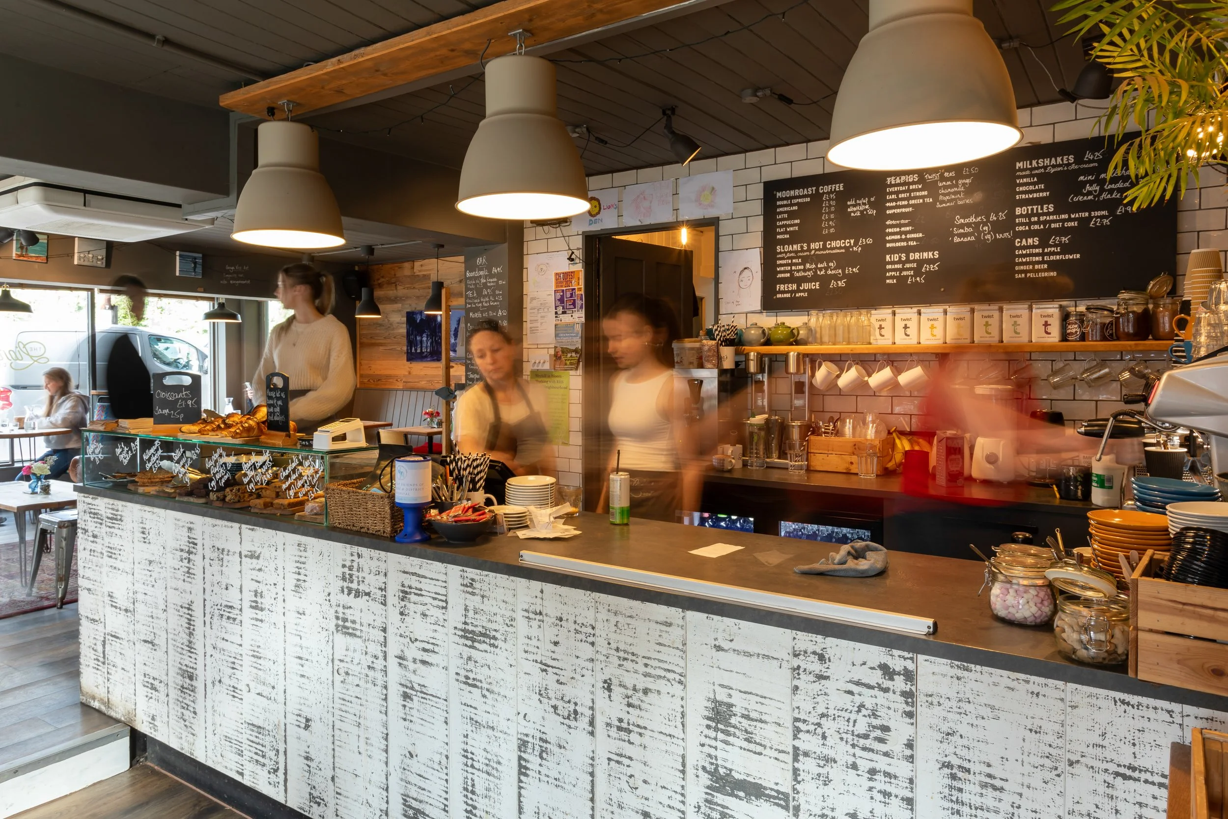 Interior of a cosy cafe with a white distressed counter, hanging pendant lights, menu boards on the wall, and staff preparing drinks.