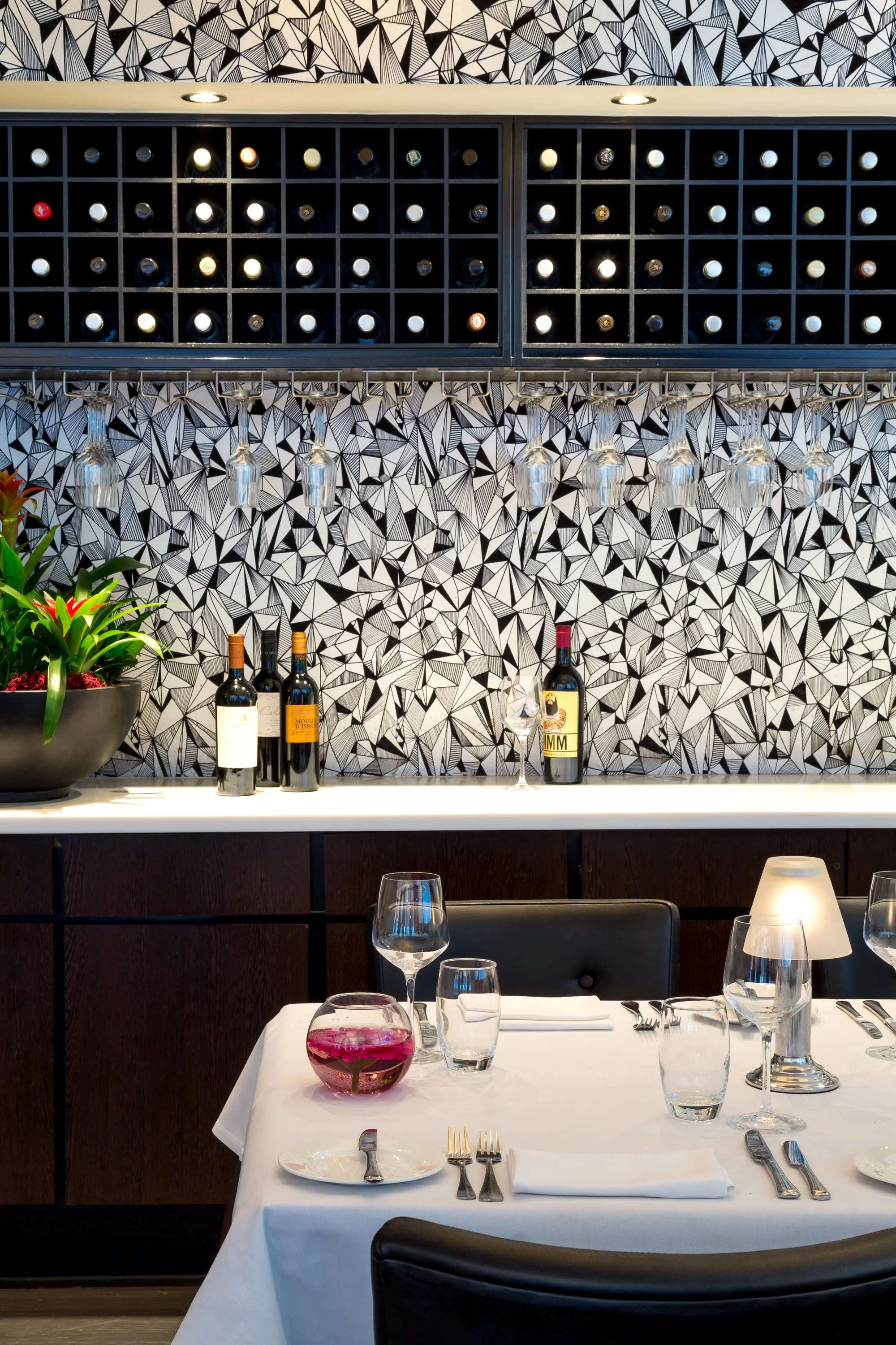 Elegant restaurant interior with a white tablecloth-dressed table, wine bottles on a patterned black and white wall, wine glasses hanging upside down, and a decorative fishbowl with pink flowers.