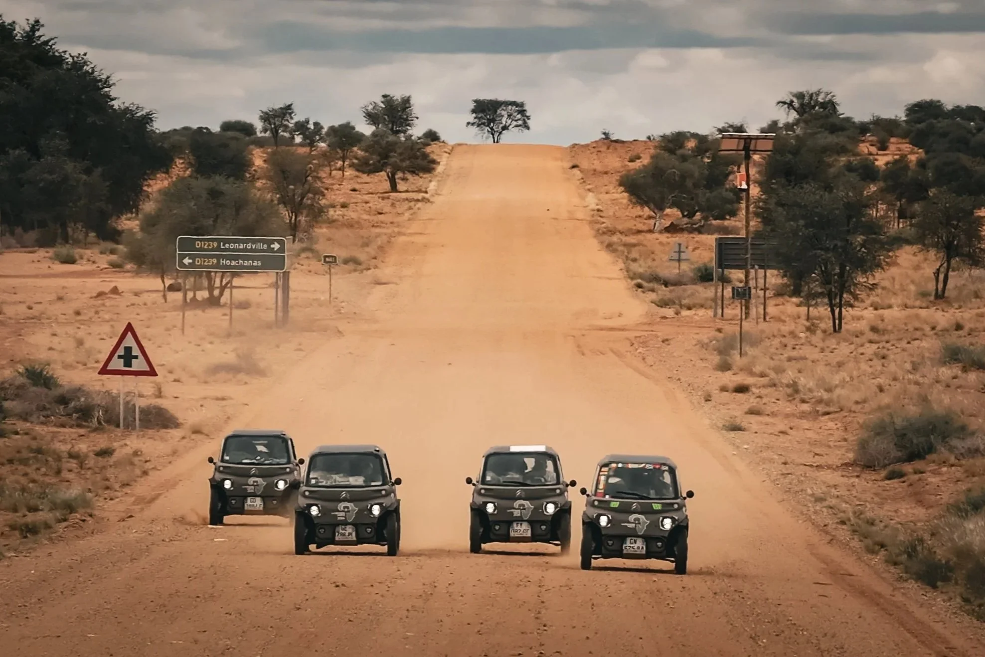 Five black cars driving on a dusty dirt road through a desert with sparse trees and cloudy skies.