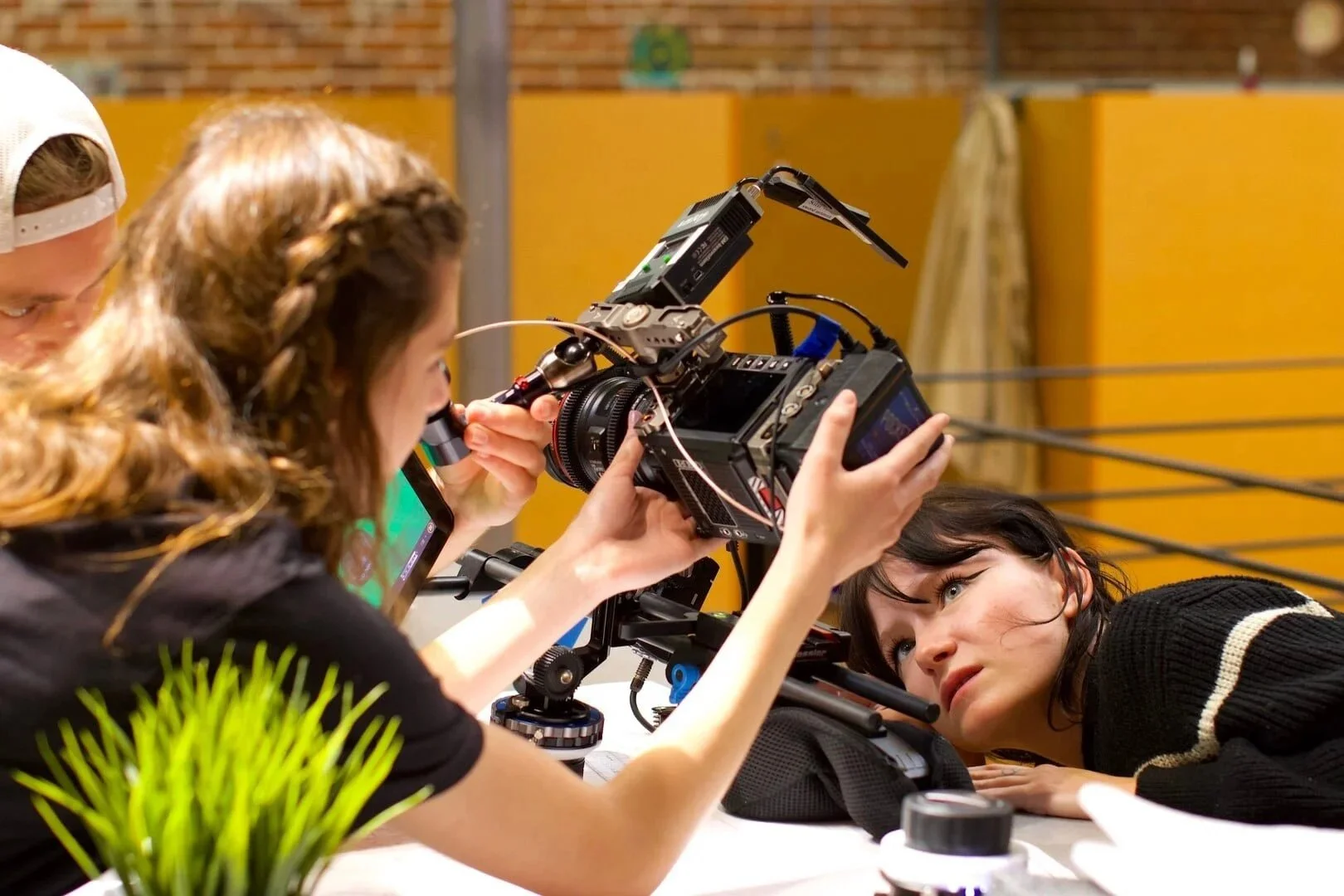 Film crew filming a woman lying on a table with a professional camera in a studio or warehouse setting.